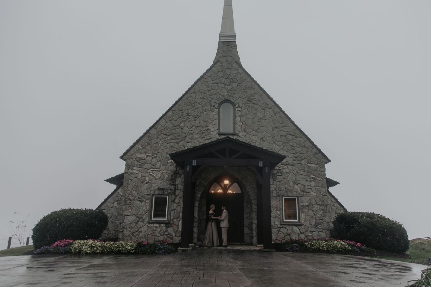 A stone chapel with a steeple stands in misty rain, with a person visible under the arched entrance adorned with warm lights.