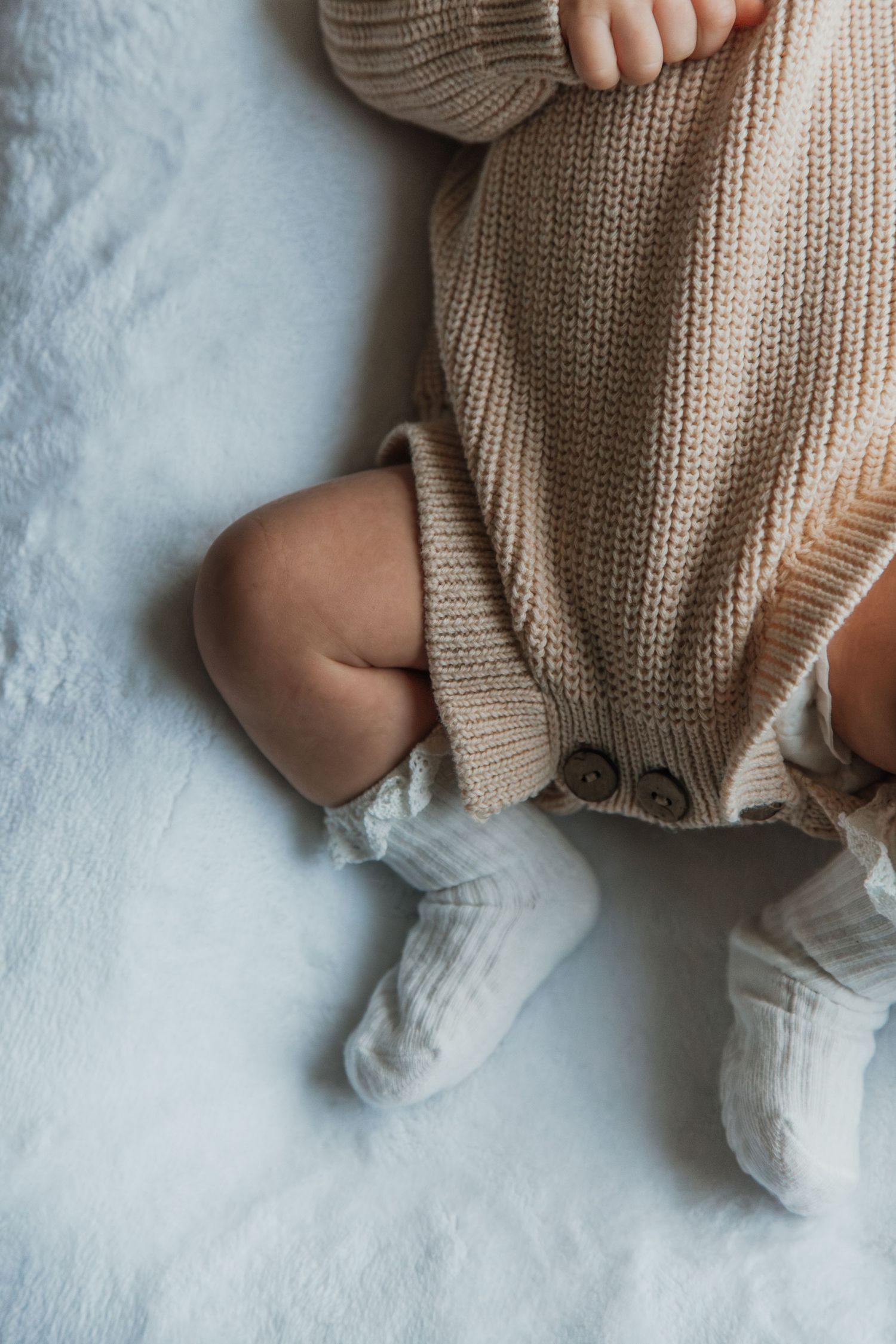 Close-up of a baby in a beige knit romper with white ribbed socks, lying on a soft light blue blanket.