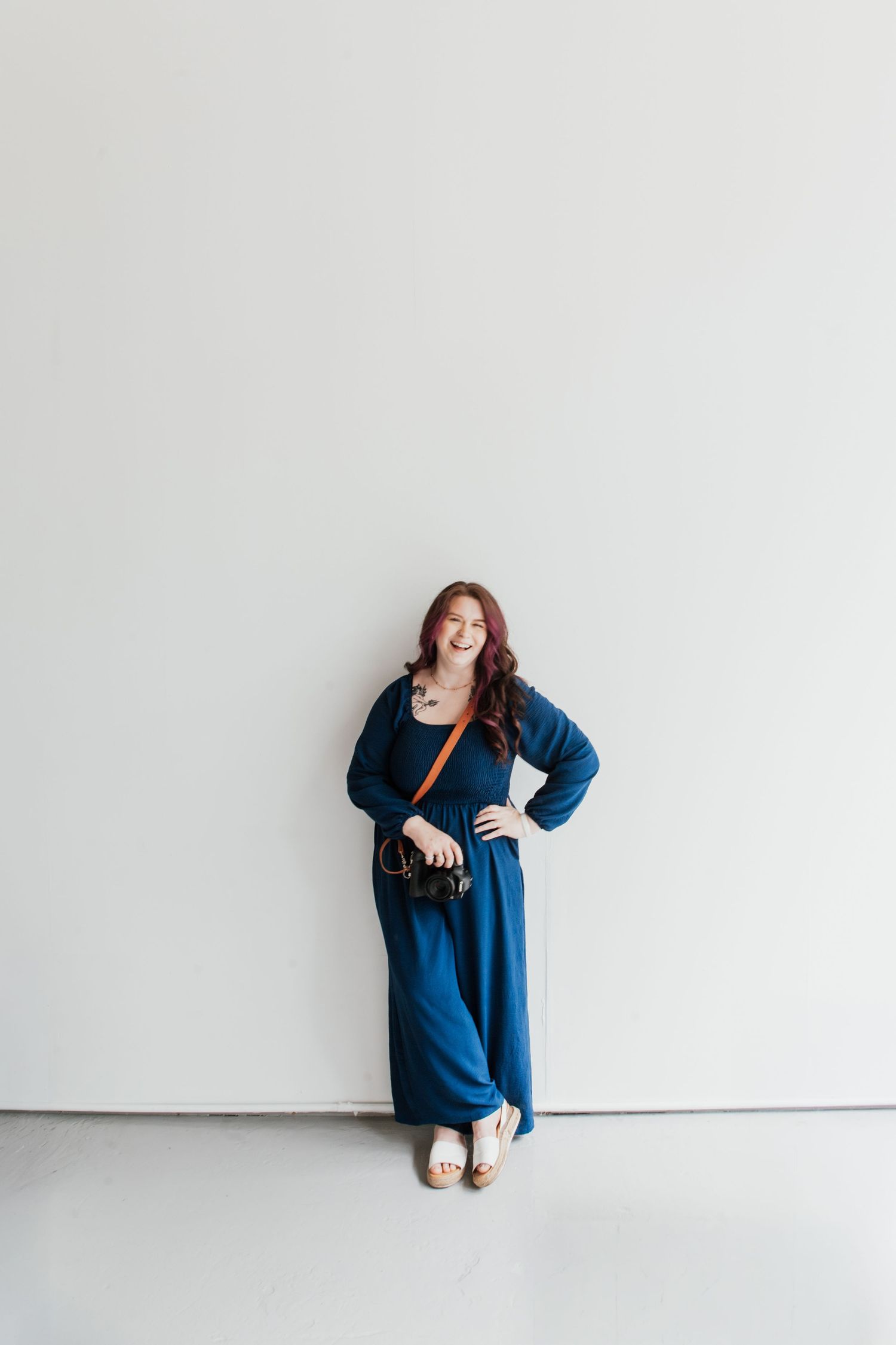 Woman in blue jumpsuit with crossbody bag smiling against white wall.