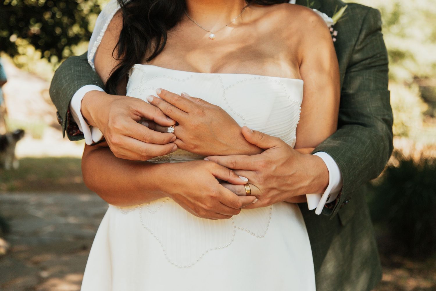 Groom in green suit embracing bride in white strapless wedding dress, both wearing wedding rings outdoors.