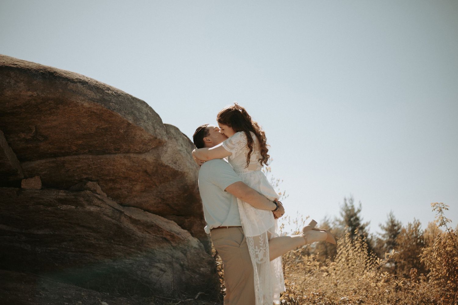 Couple embracing and kissing near a large rock formation in a sunny outdoor setting with dry golden grass.
