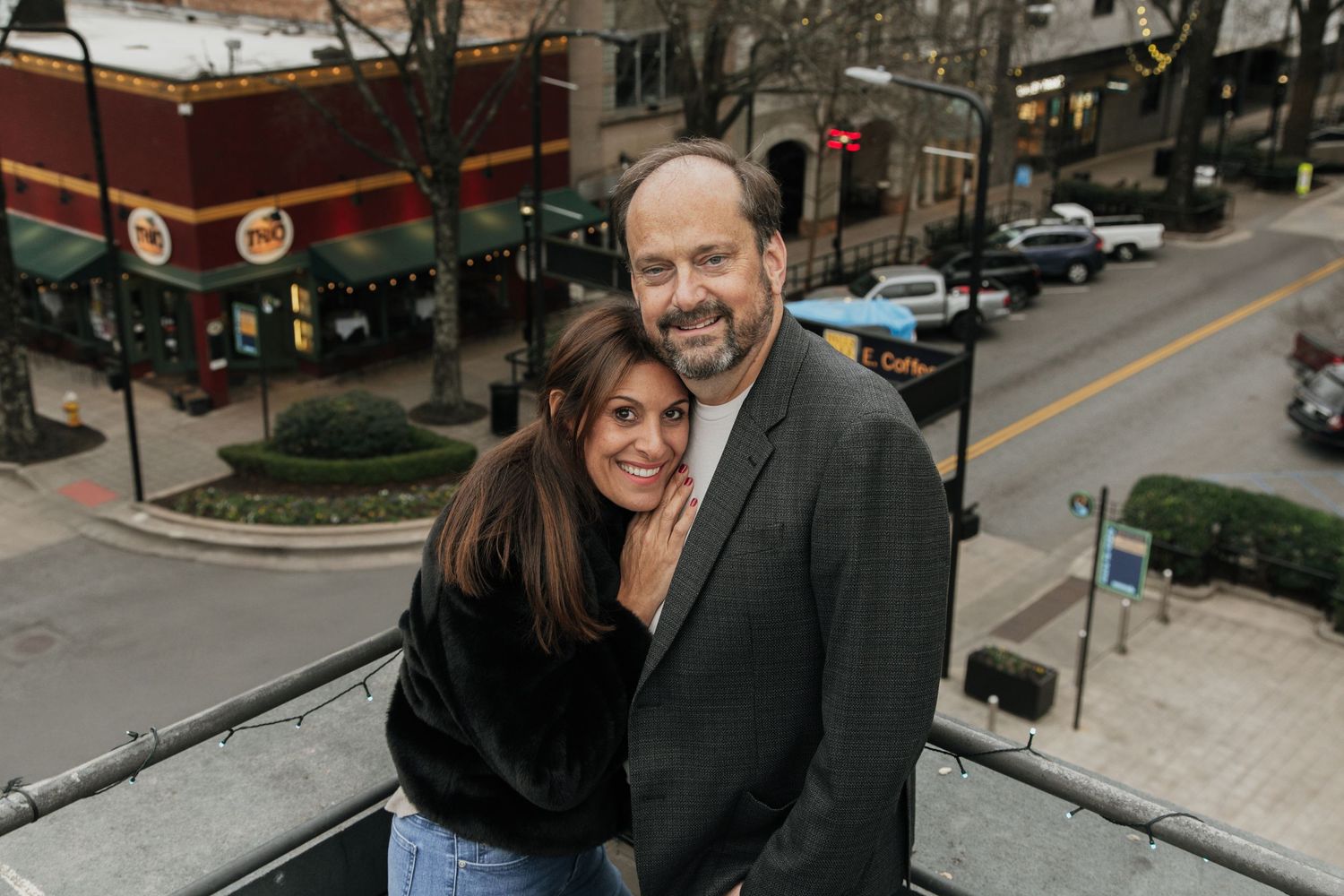 Couple smiling and embracing on an outdoor balcony overlooking a charming urban street with shops below.