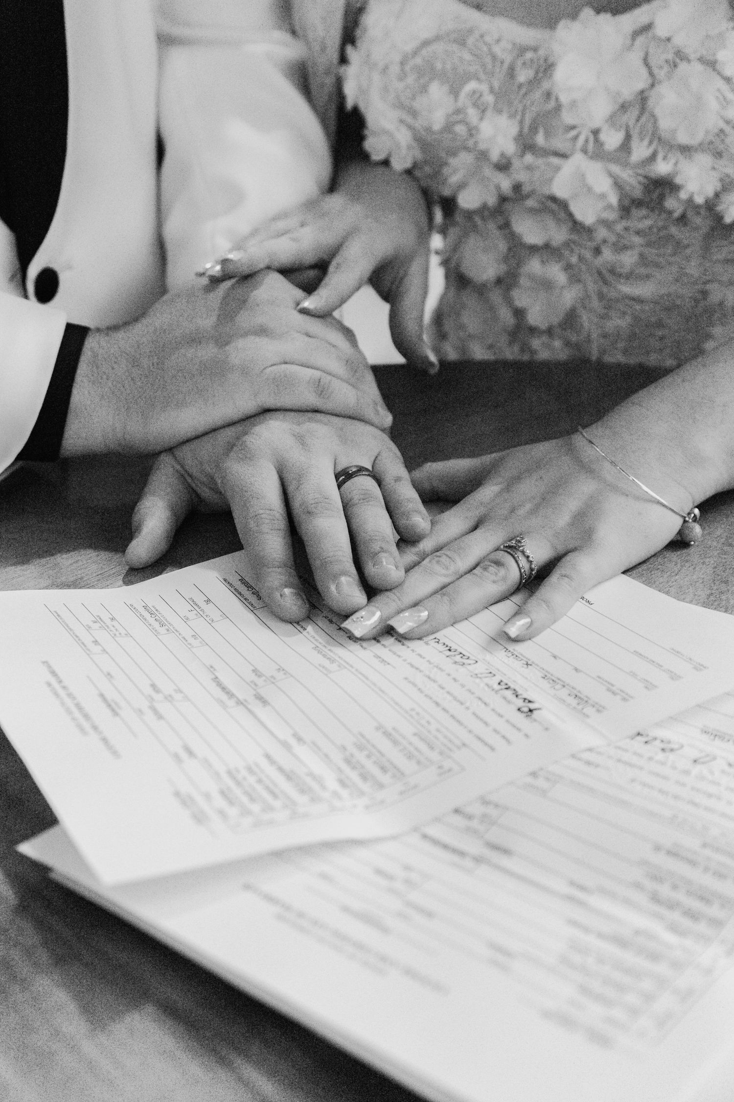 Black and white close-up of newlyweds hands with wedding rings resting on signed marriage certificate document.