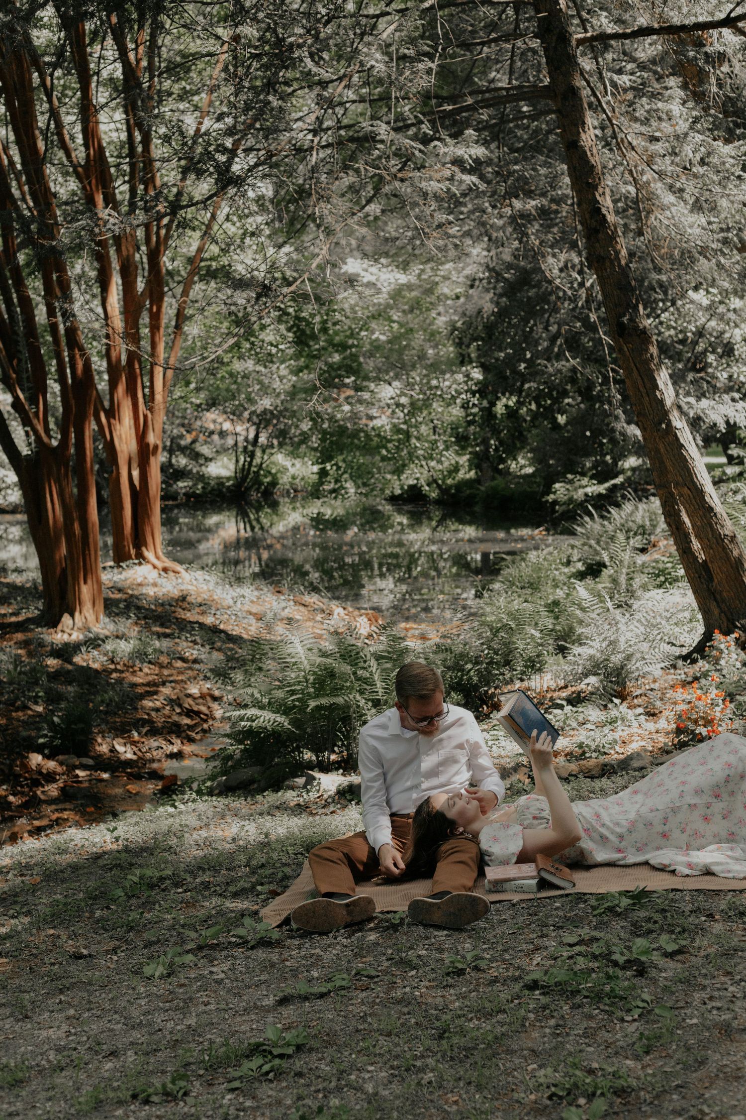 Couple relaxing on a blanket by a tranquil forest pond, reading together in dappled sunlight. kilgore lewis house greenville sc