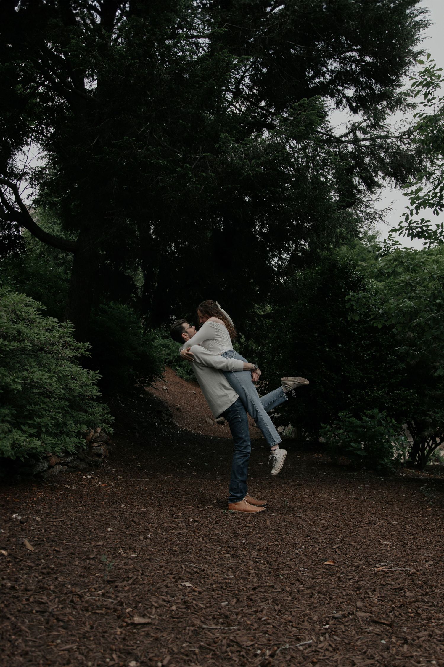 Man lifting woman playfully in a moody dark forest during a casual outdoor couples photo session. rock quarry garden greenville sc