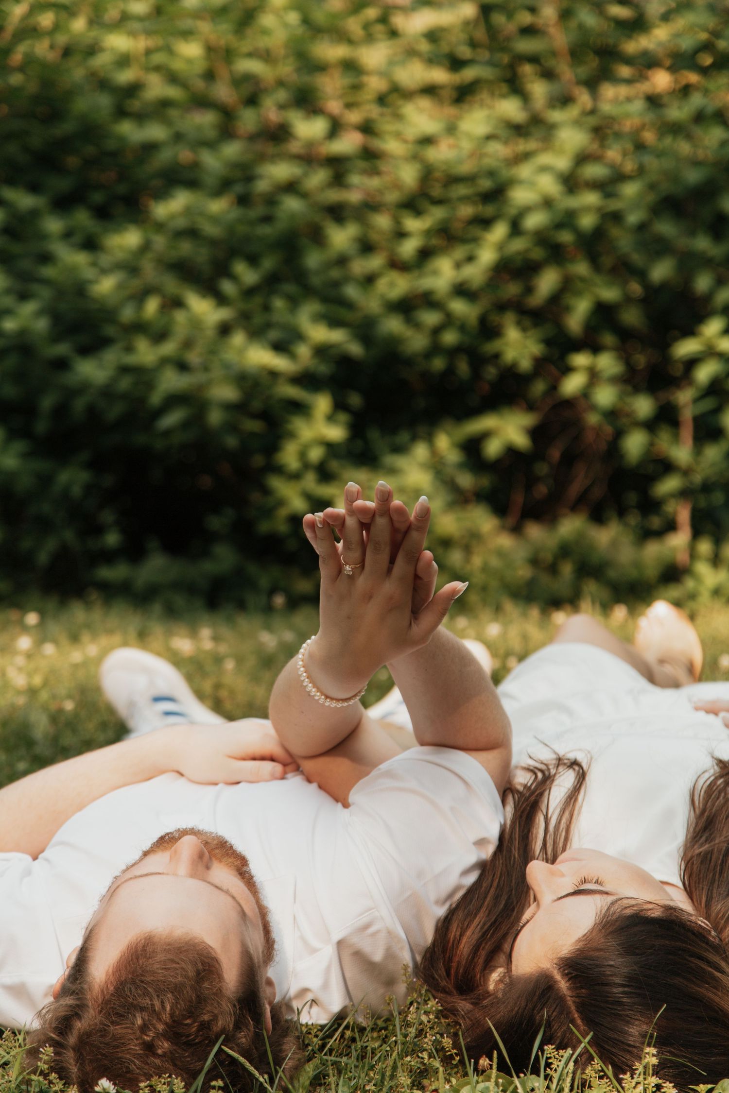 Couple lying on grass holding hands up, woman wearing pearl bracelet and engagement ring.