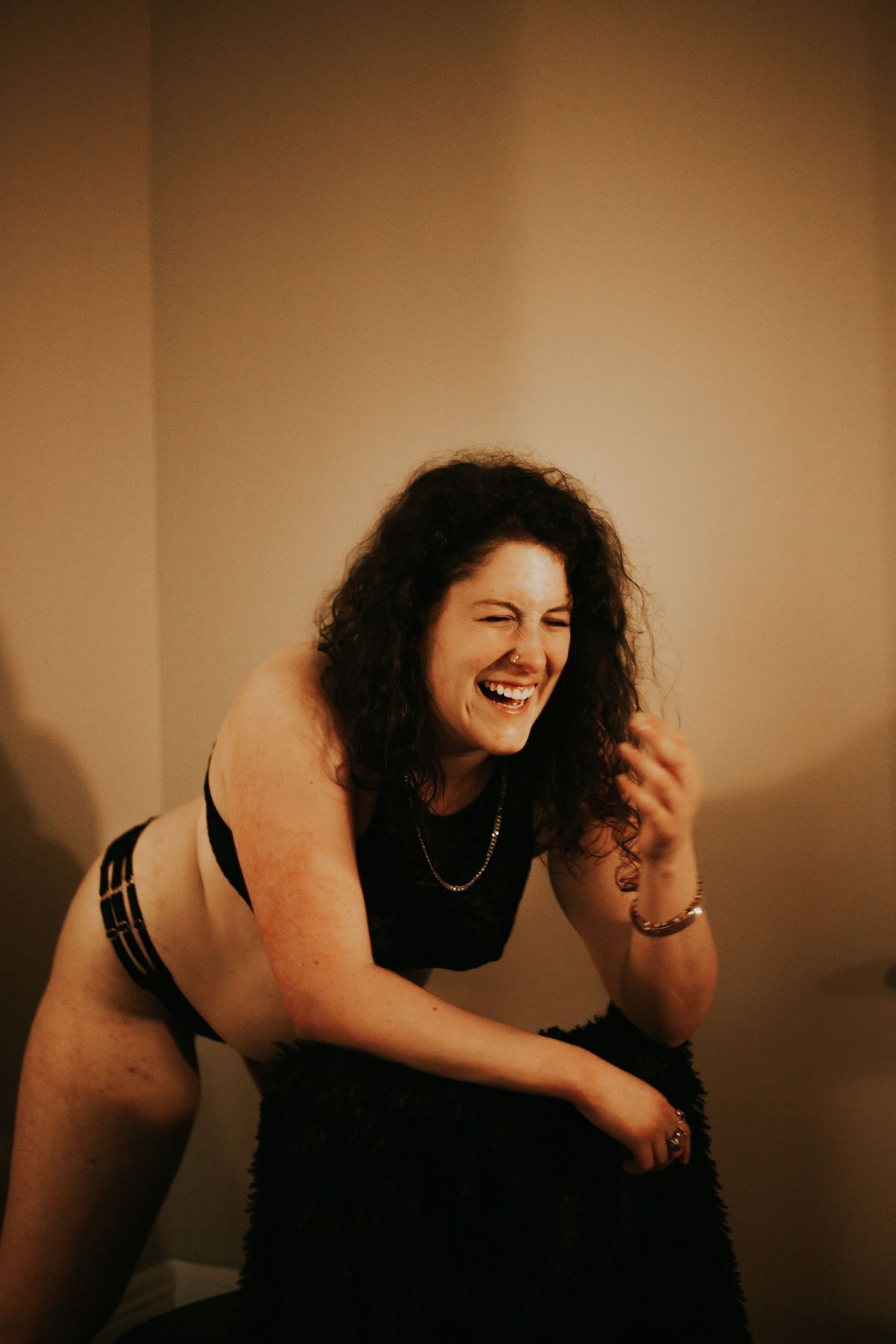A curly-haired woman laughs joyfully in black lingerie during a boudoir photography session.