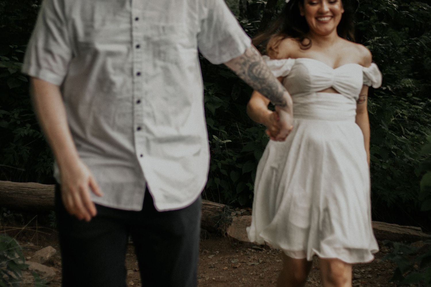 Tattooed groom leads bride in white dress along a forest path.