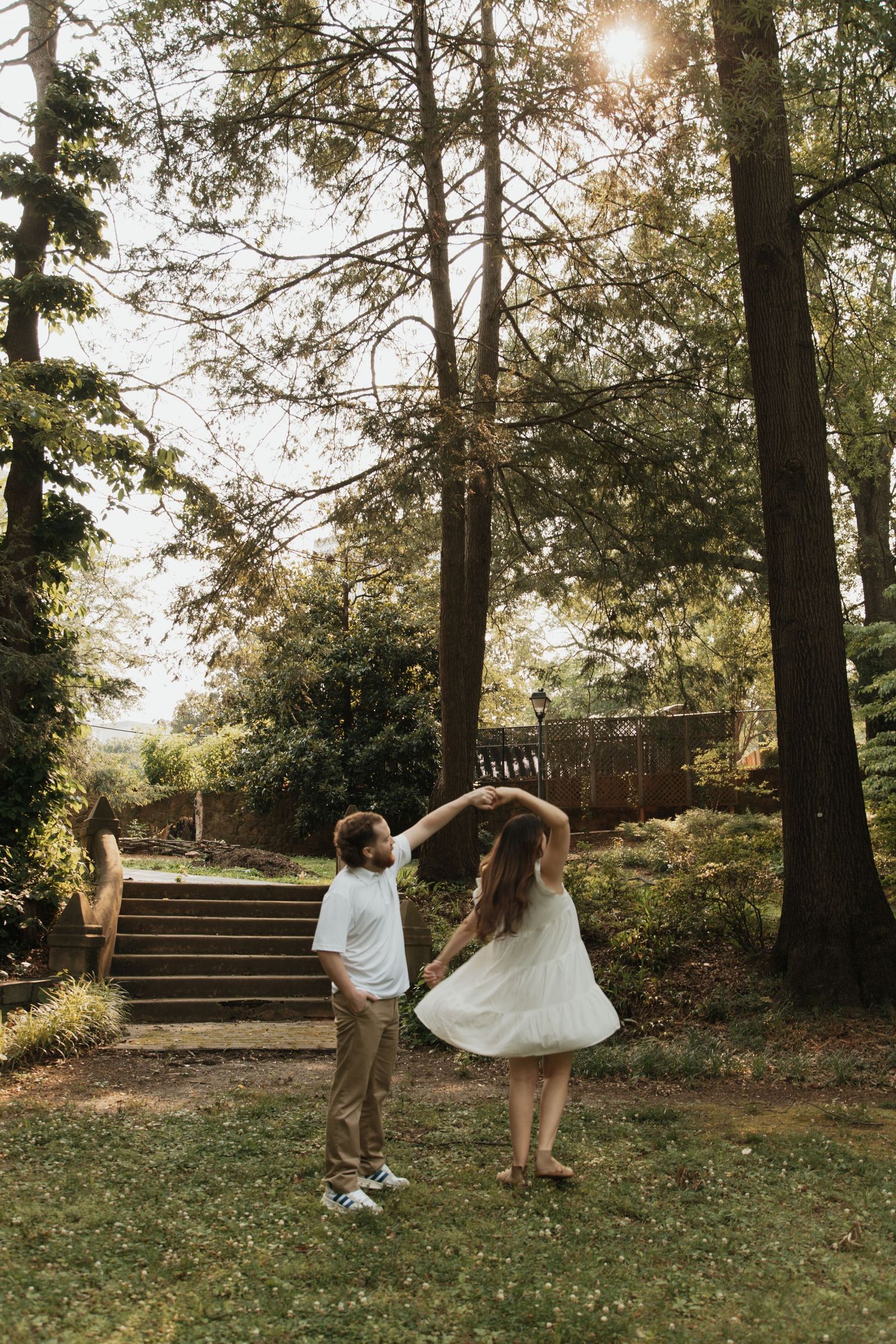 Couple dancing joyfully in sunlit garden with tall trees and stone steps in background. kilgore lewis house and garden greenville sc