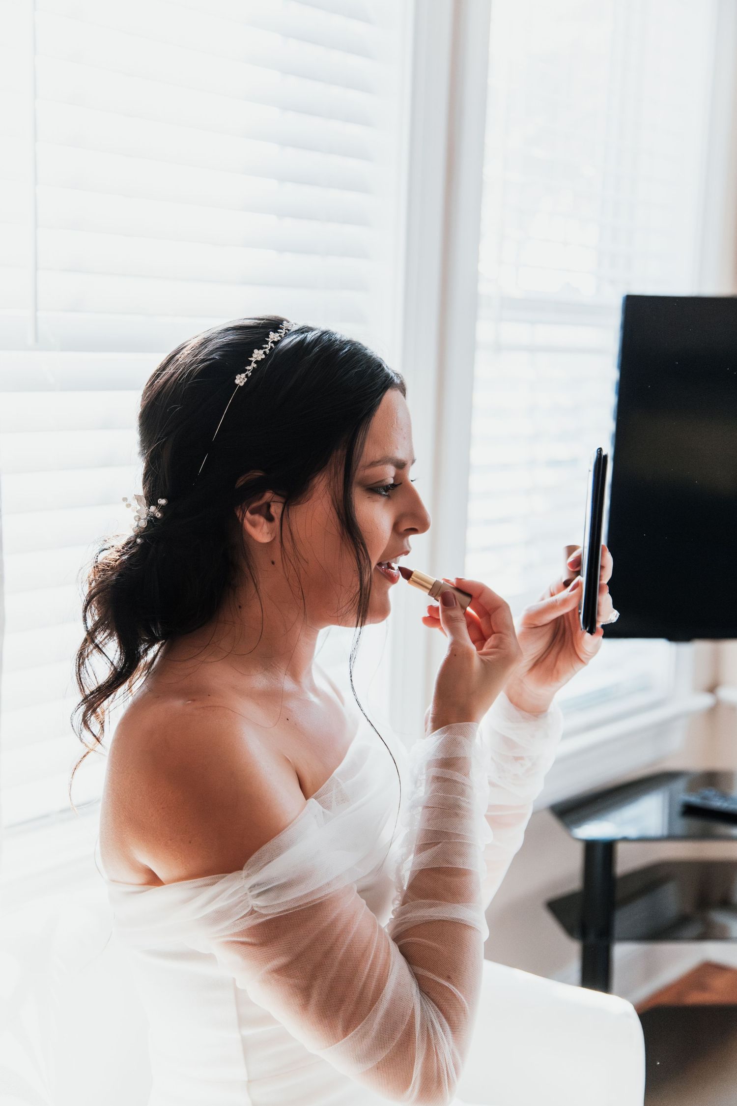 Bride applying lipstick using a handheld mirror while wearing an off-shoulder white gown and floral hair accessory.