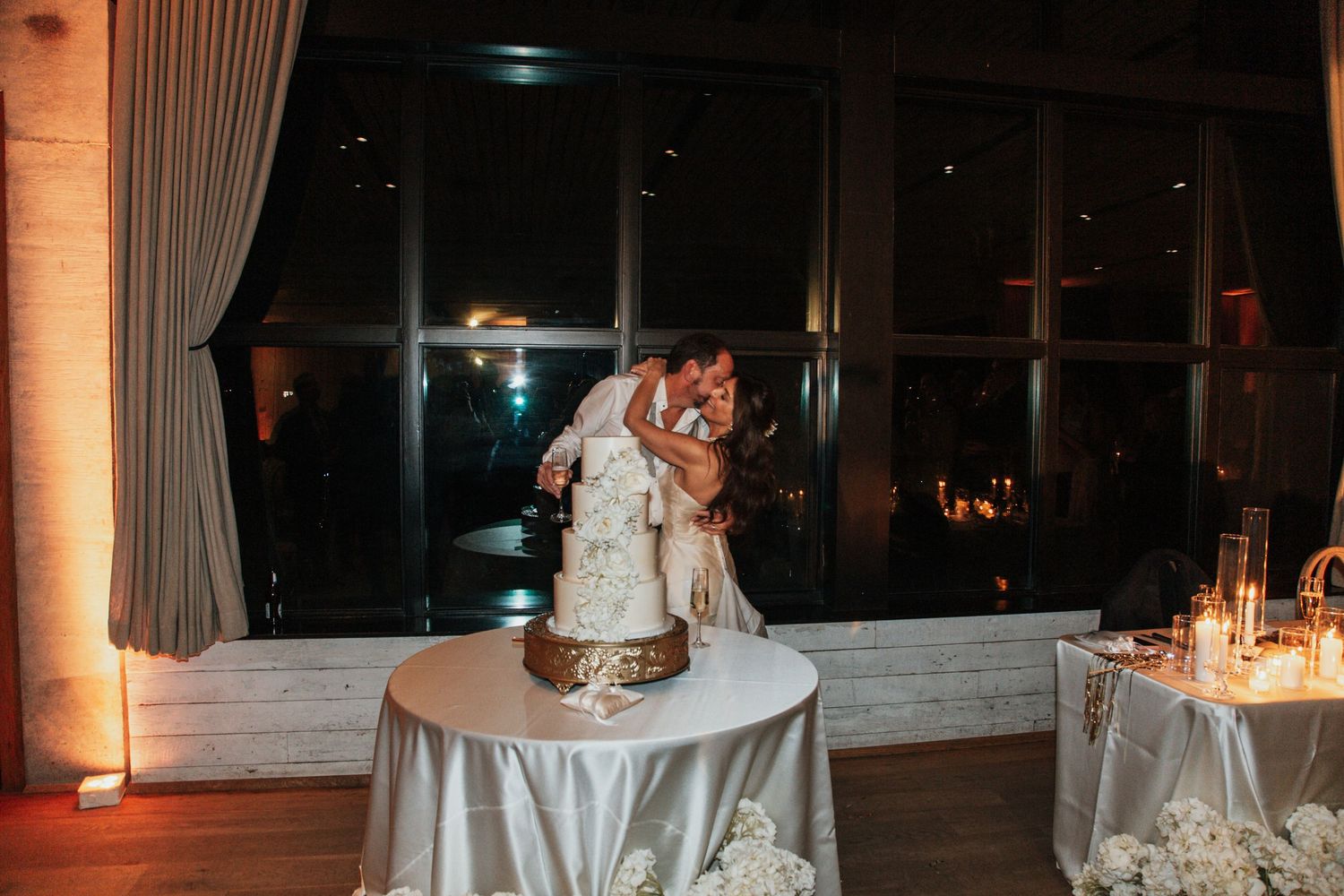 Couple sharing romantic kiss beside white wedding cake at dimly lit elegant reception with warm ambient lighting. middleton place sc