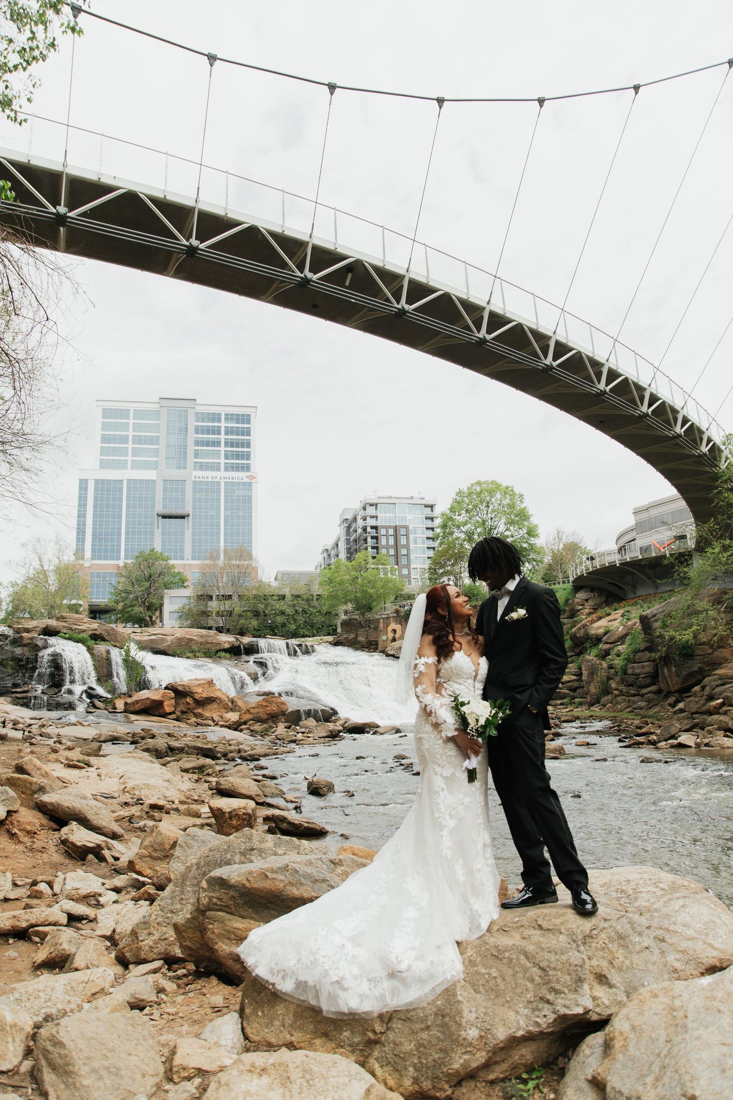 Bride and groom kiss on rocky riverbank beneath a suspension bridge with waterfall backdrop.  falls park greenville sc