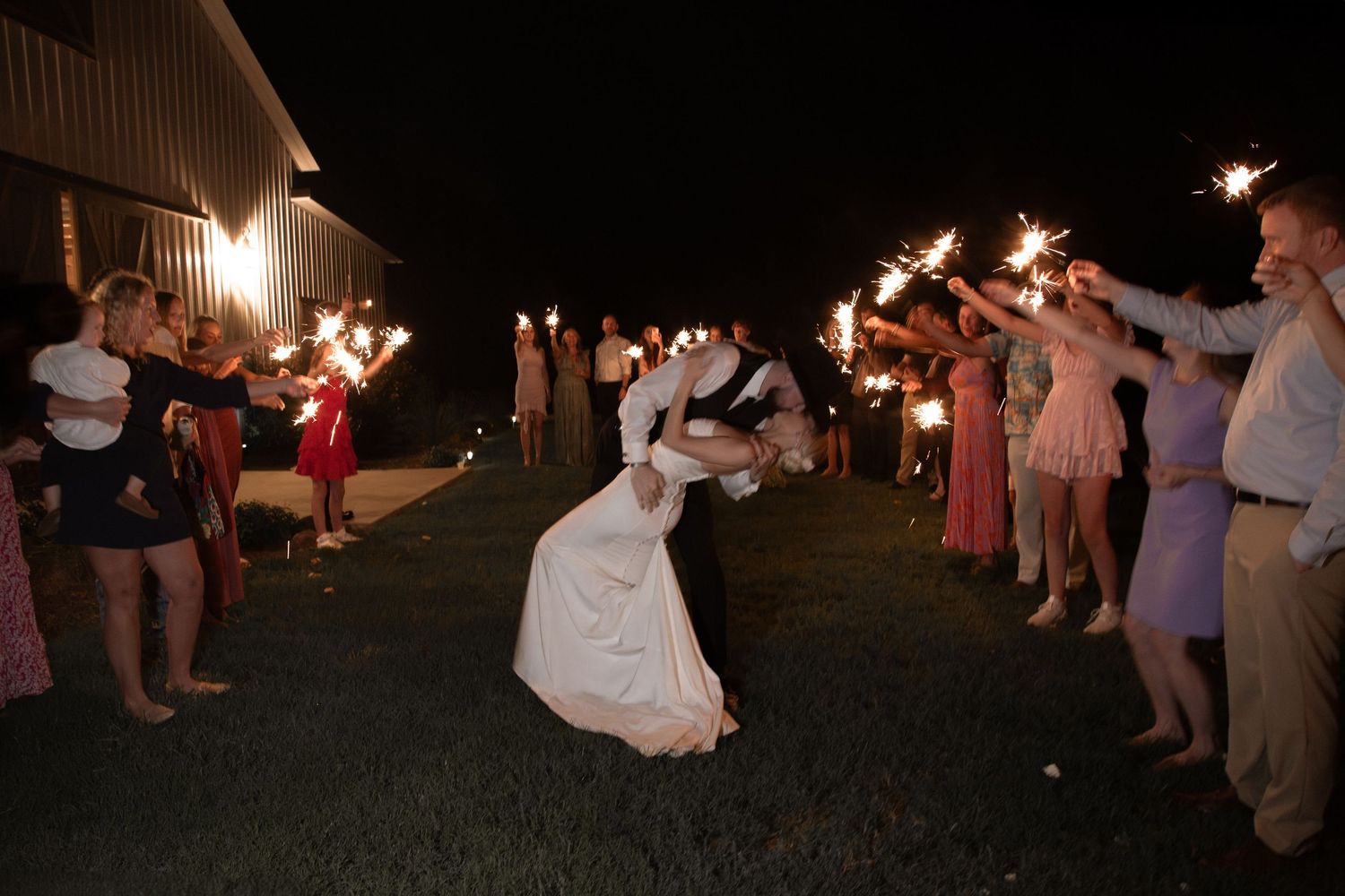 Bride and groom kiss during sparkler send-off exit surrounded by cheering wedding guests at night. Roger's Farm Buffalo SC.