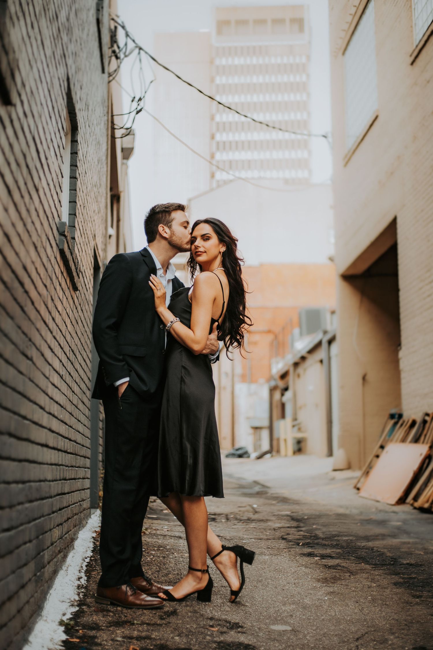 Couple in formal attire sharing a romantic kiss in an urban alley, woman in black dress, man in dark suit.