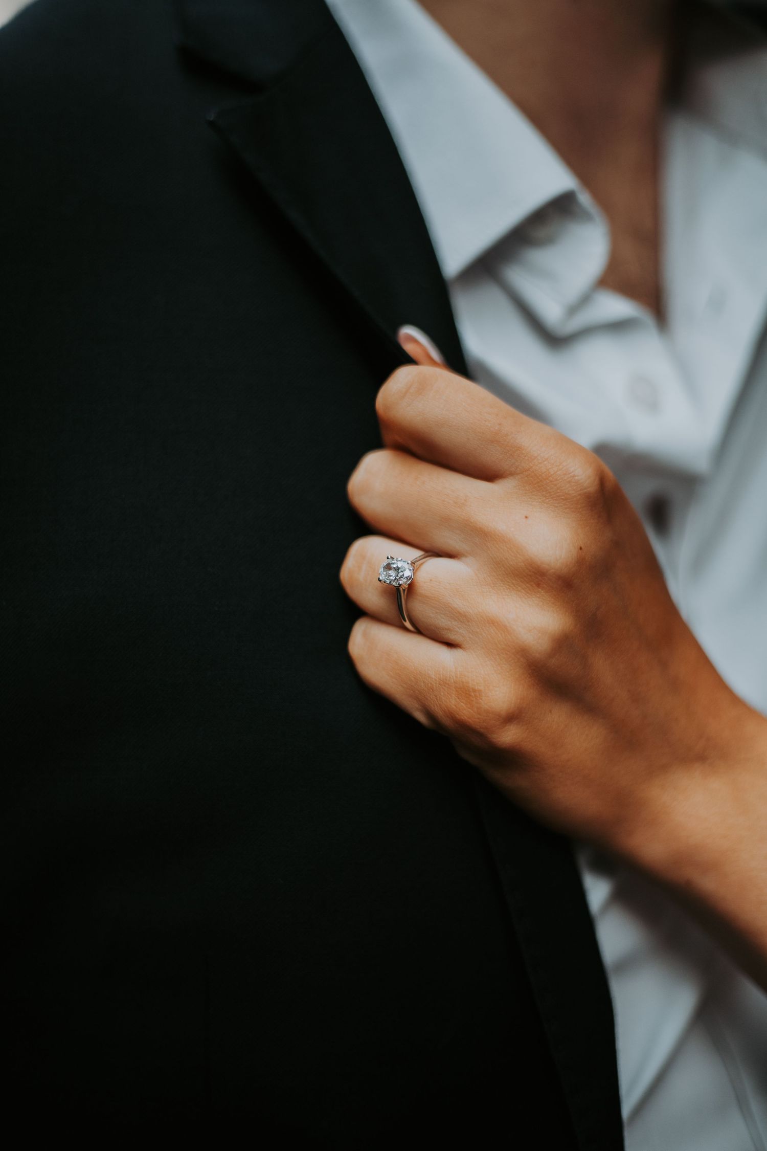 Close-up of an engagement ring on a womans hand resting on a black suit jacket.