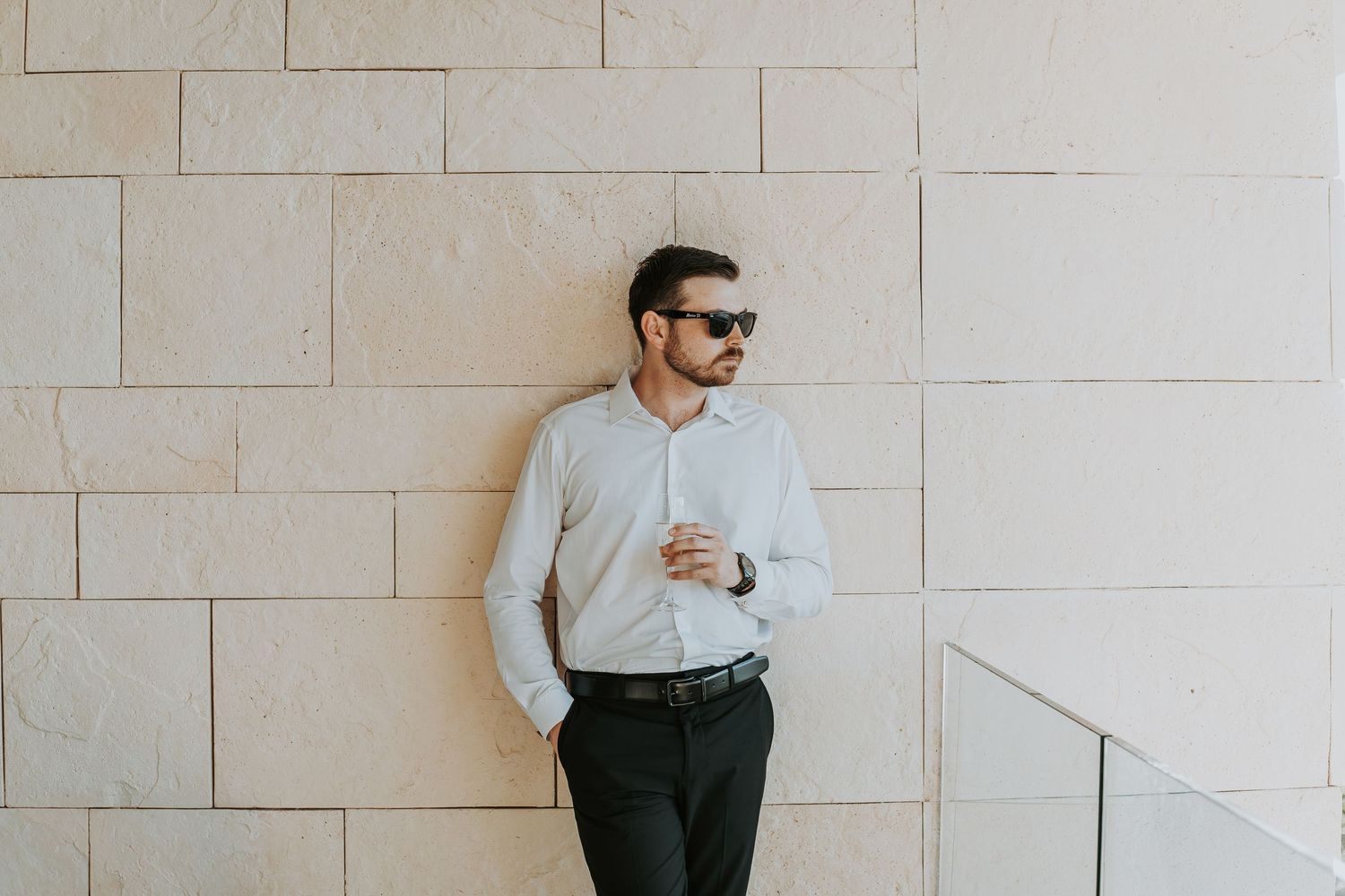 Stylish groom in white shirt and sunglasses leans against stone wall before wedding. - riviera maya resort (cancun, Mexico)