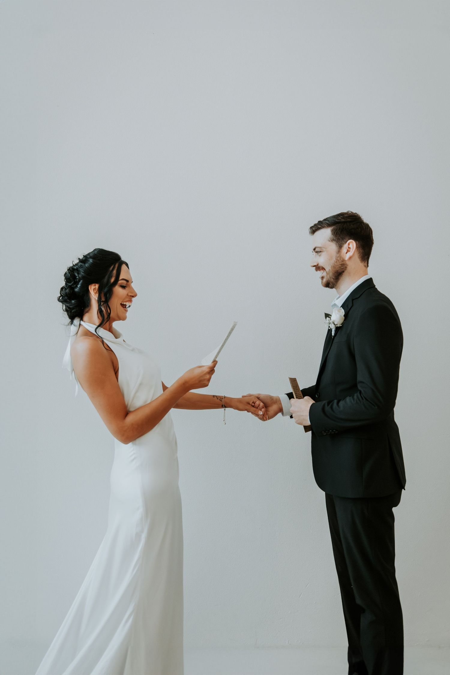 Smiling bride in white gown reads vows while holding hands with groom in black tuxedo against white backdrop.