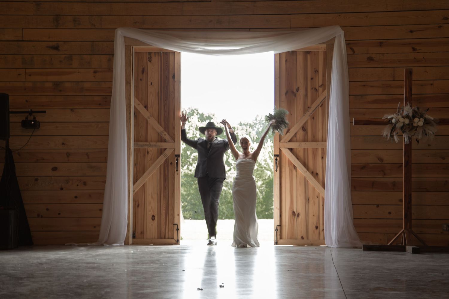 Bride and groom raise arms in celebration at rustic barn wedding venue with draped white doors. Roger's Farm Buffalo SC.