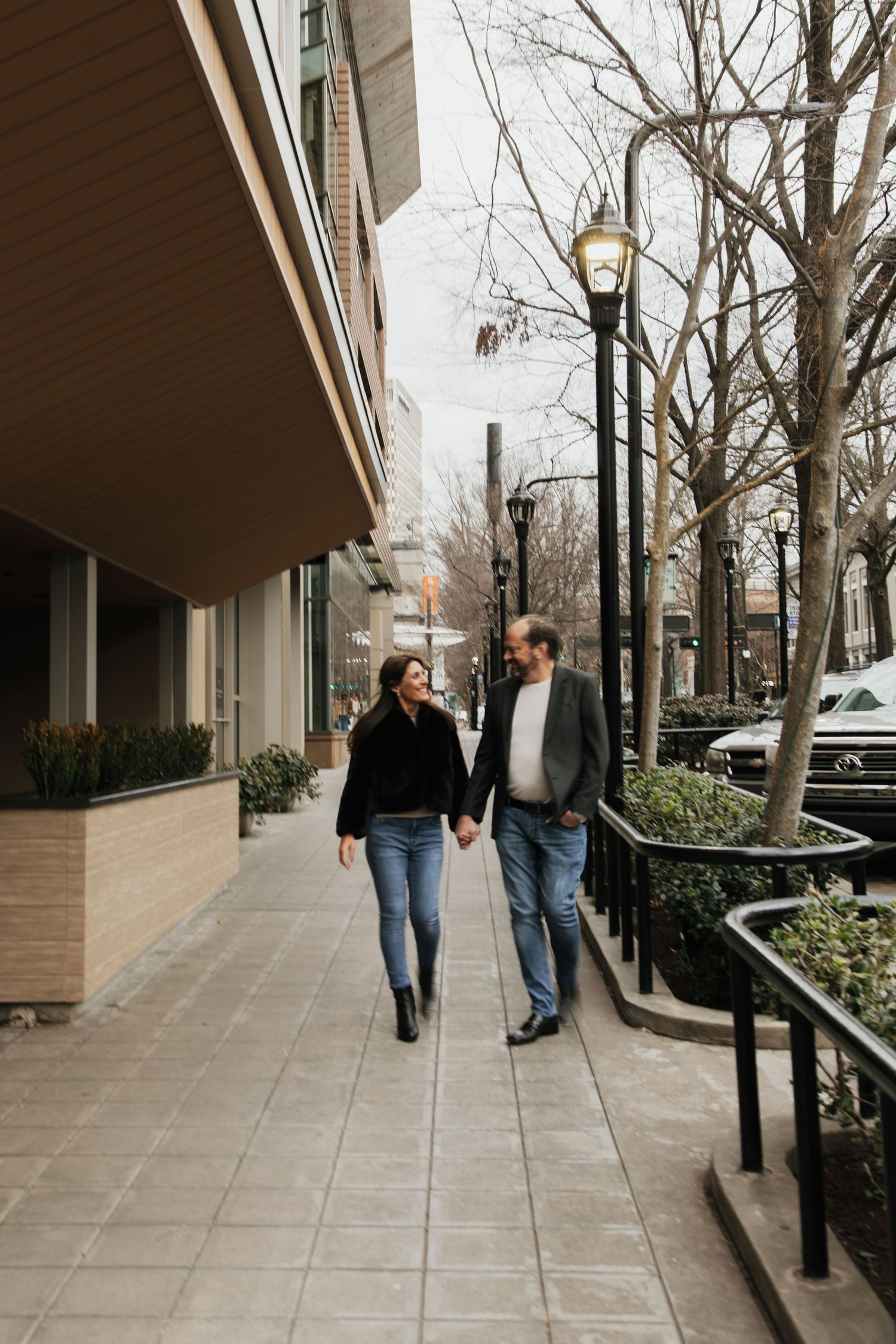 Couple holding hands and smiling while walking along an urban sidewalk lined with lamp posts and bare trees.