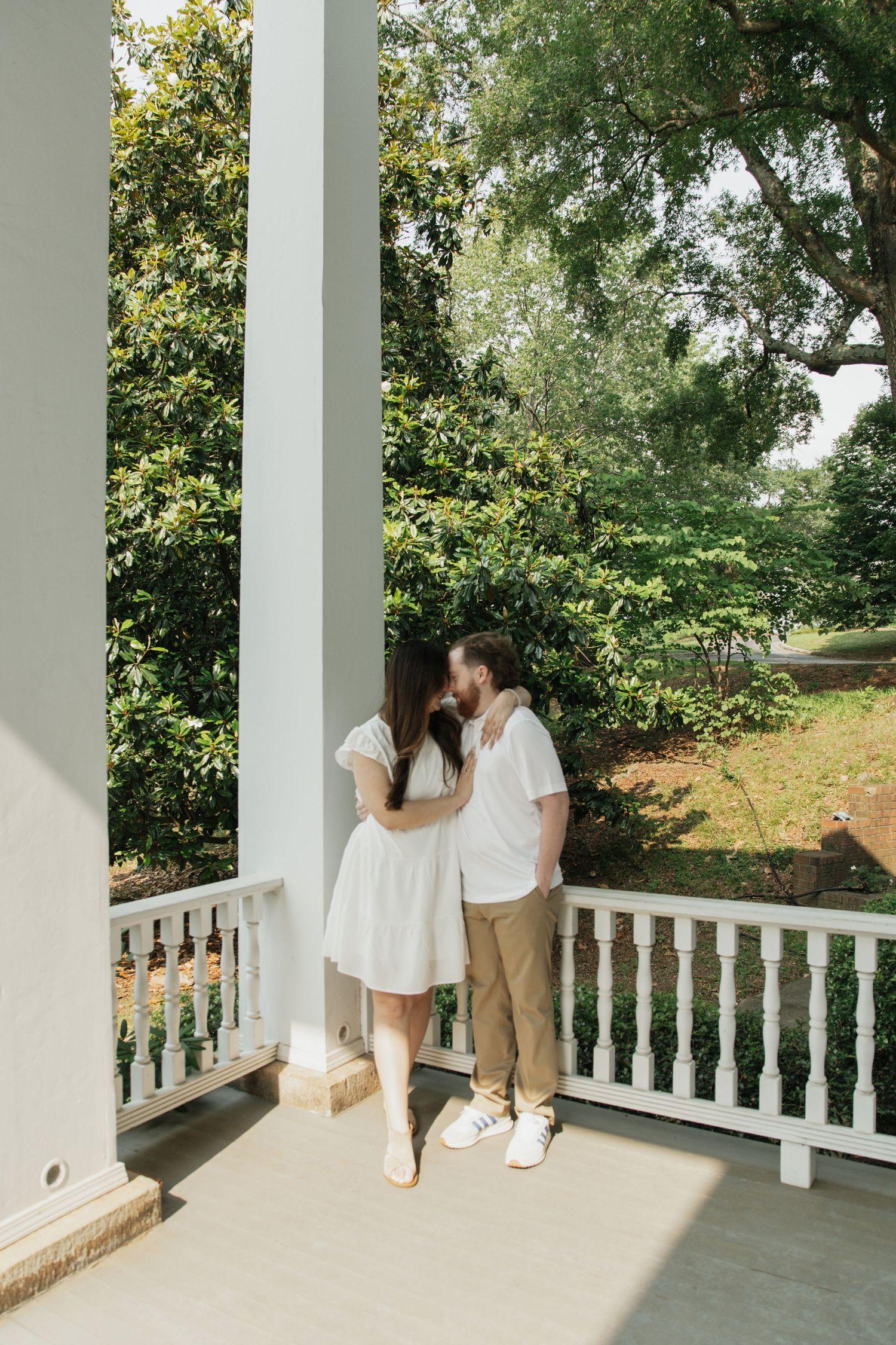 Couple sharing a romantic kiss on a white Southern-style porch surrounded by lush green trees and garden.