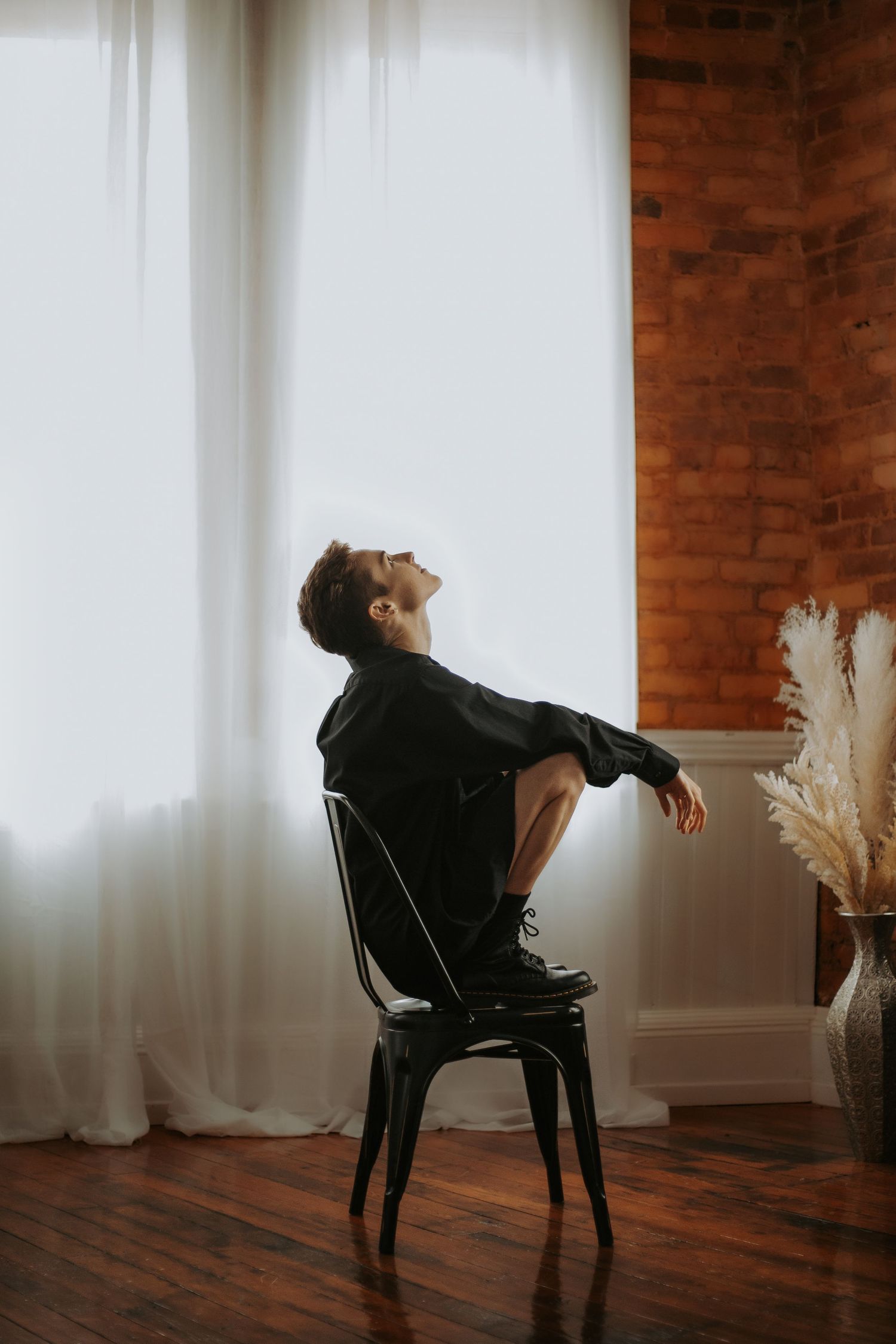 Person in black outfit sitting on a chair, leaning back with eyes closed near a bright window and brick wall.
