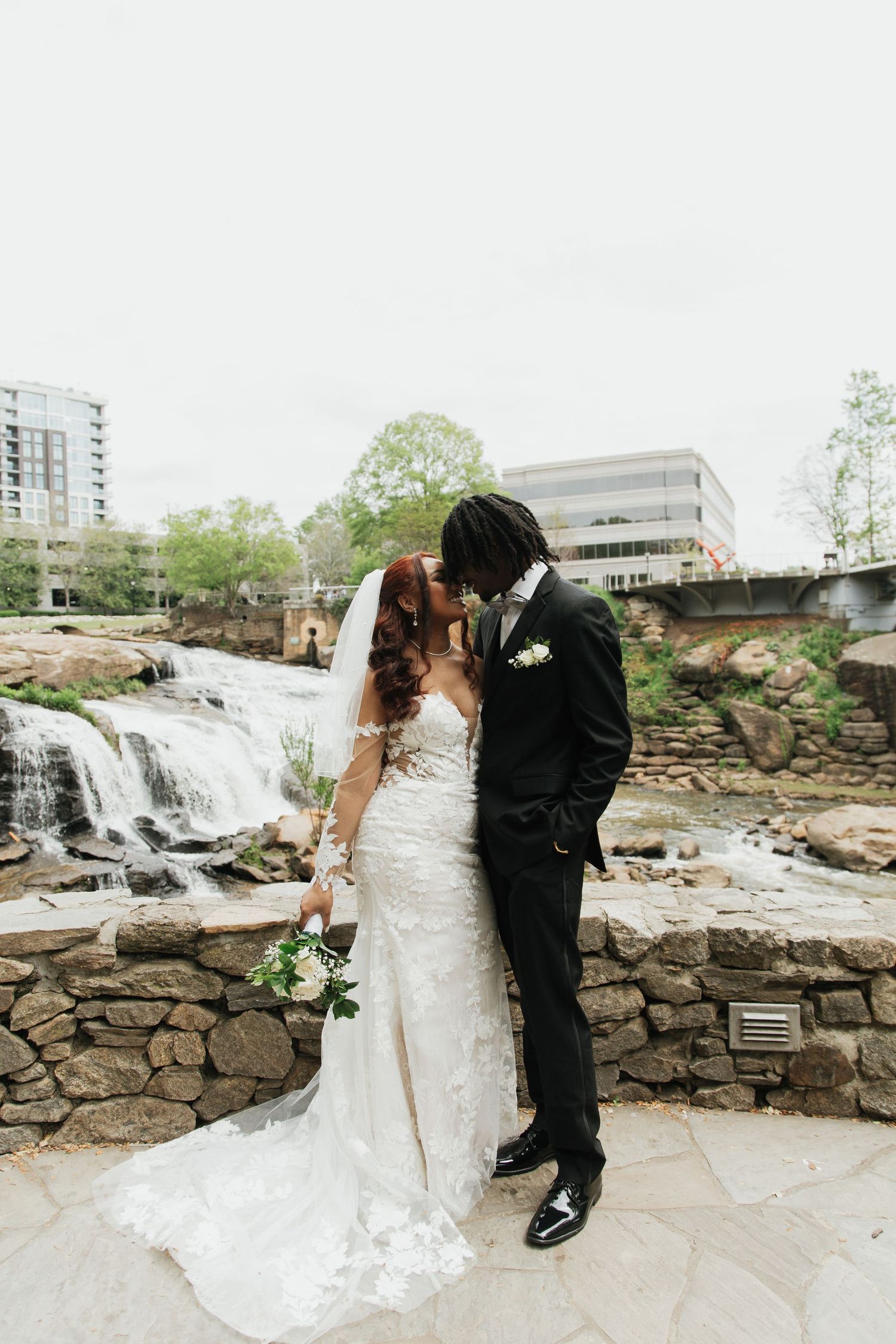 Bride and groom share a romantic kiss in front of a waterfall, bride in lace gown holding white bouquet.