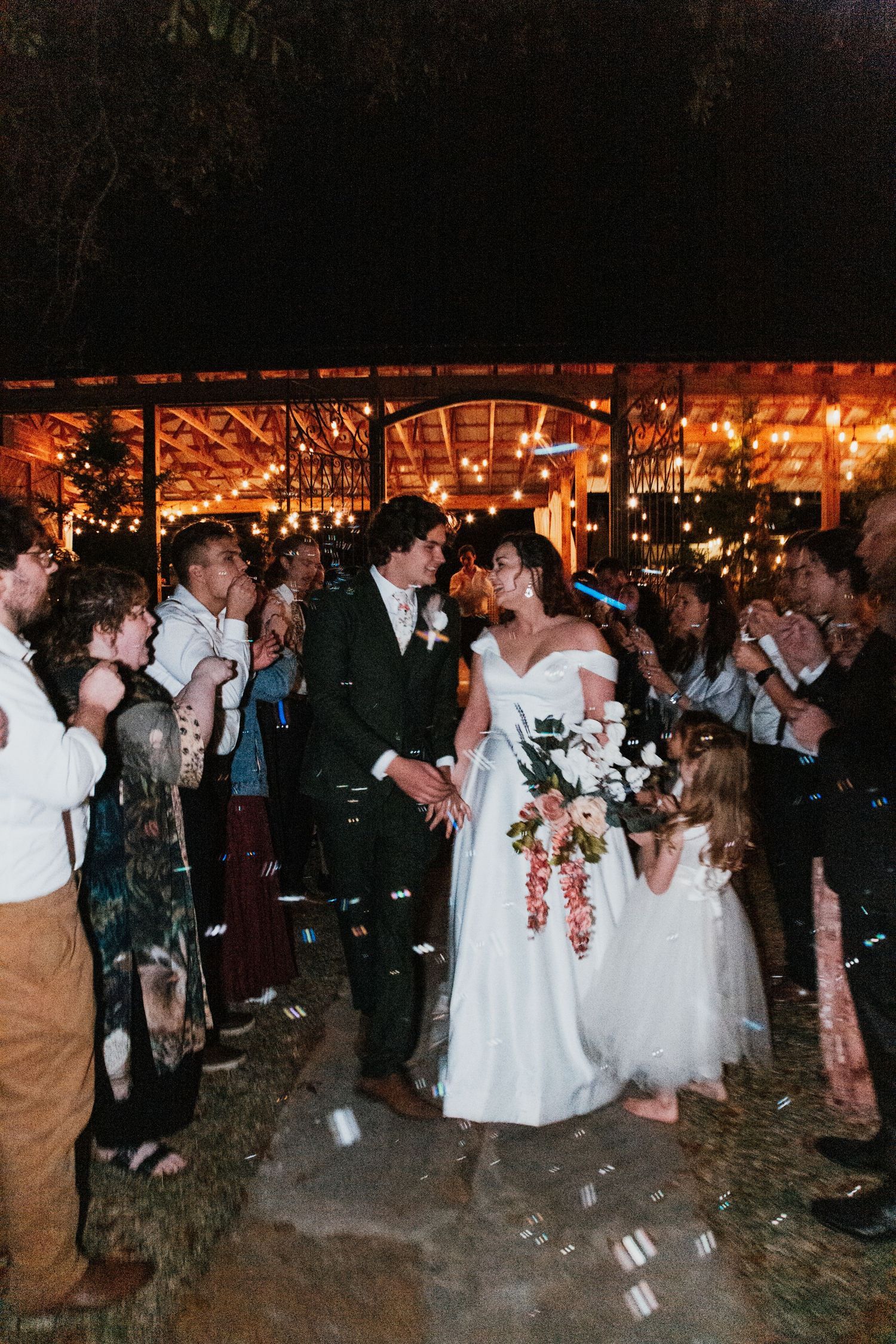Bride and groom walk through cheering guests at night wedding reception with warm string lights glowing in background.