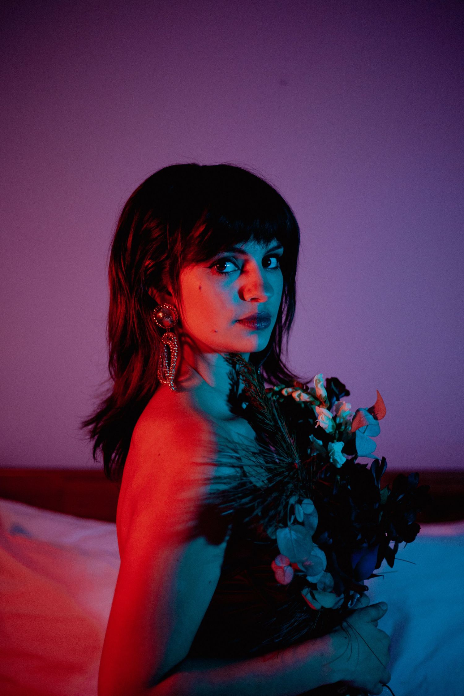 A dark-haired woman holds a bouquet of flowers under dramatic red and blue neon lighting in a bedroom.