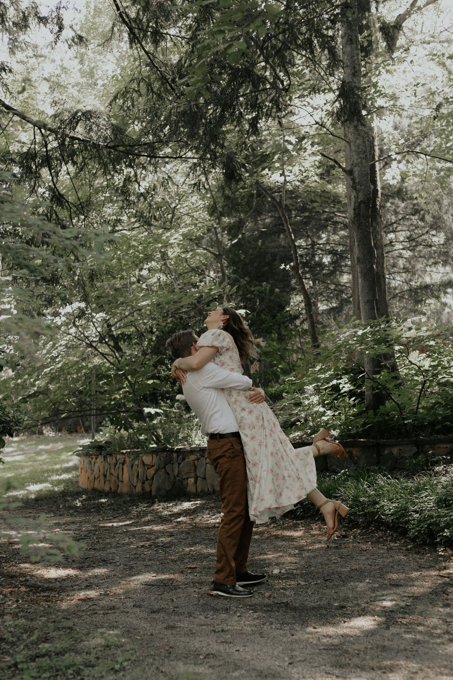 Man joyfully lifting woman in floral dress in a lush green forest during an outdoor engagement session. kilgore lewis house greenville sc