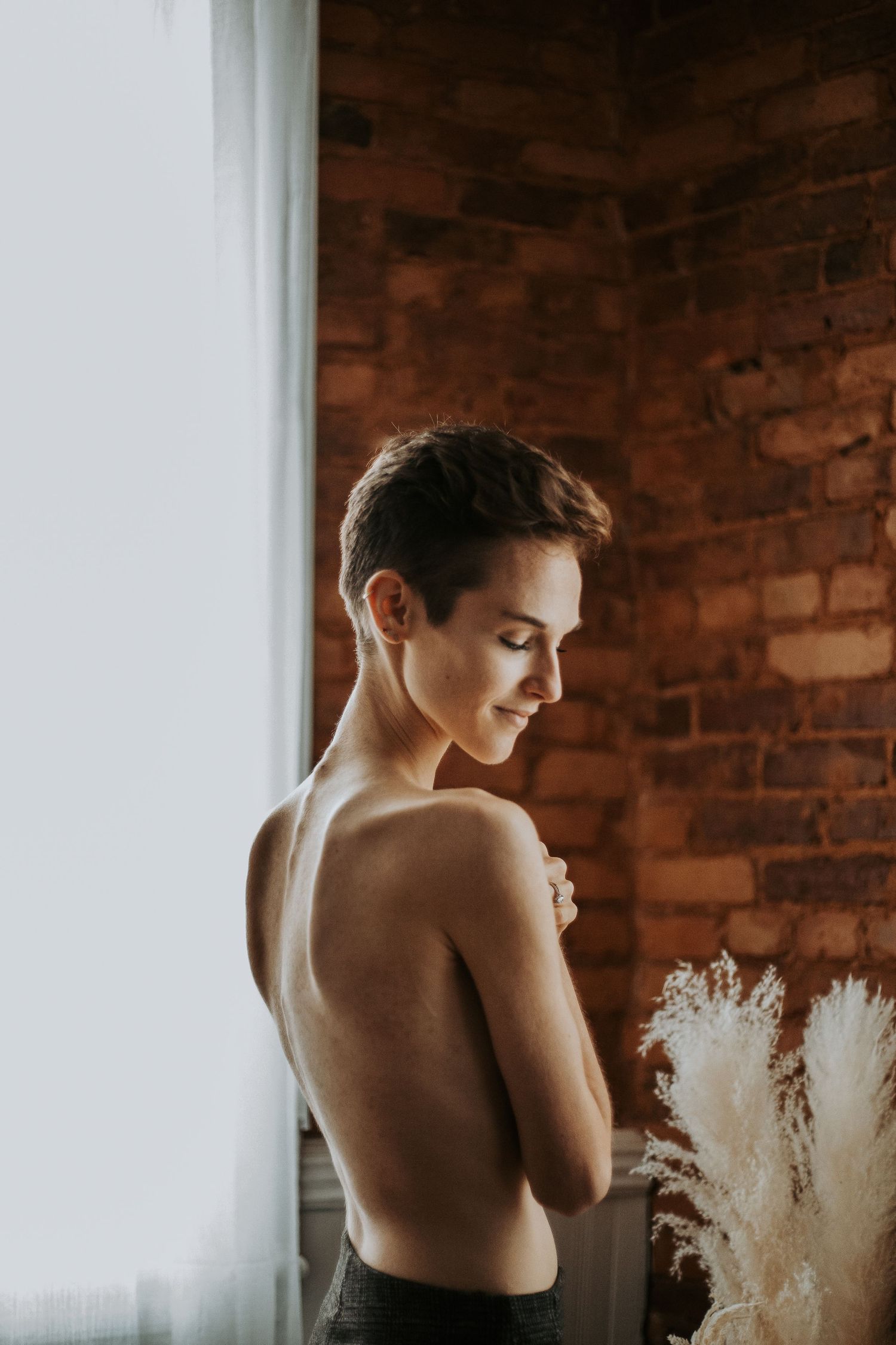 A young woman with short hair poses topless near a window with a brick wall background and pampas grass.