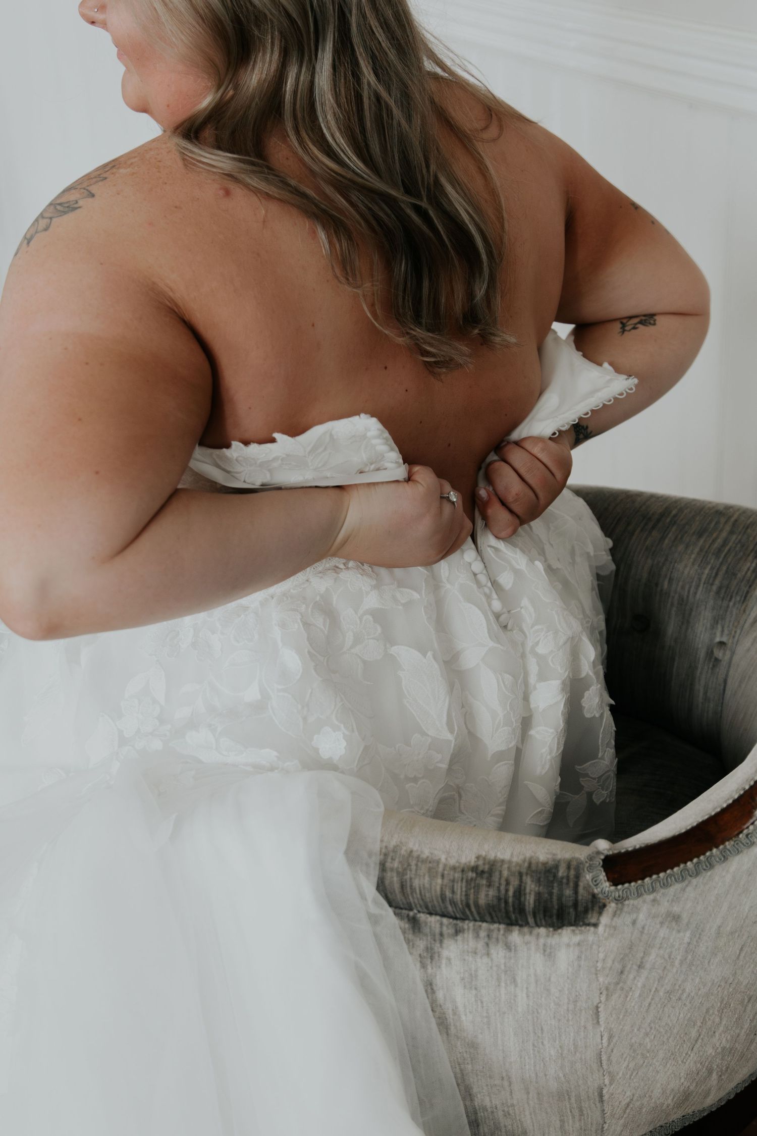 Bride in a white strapless wedding gown adjusting her dress from behind, showcasing tattoos and flowing skirt.