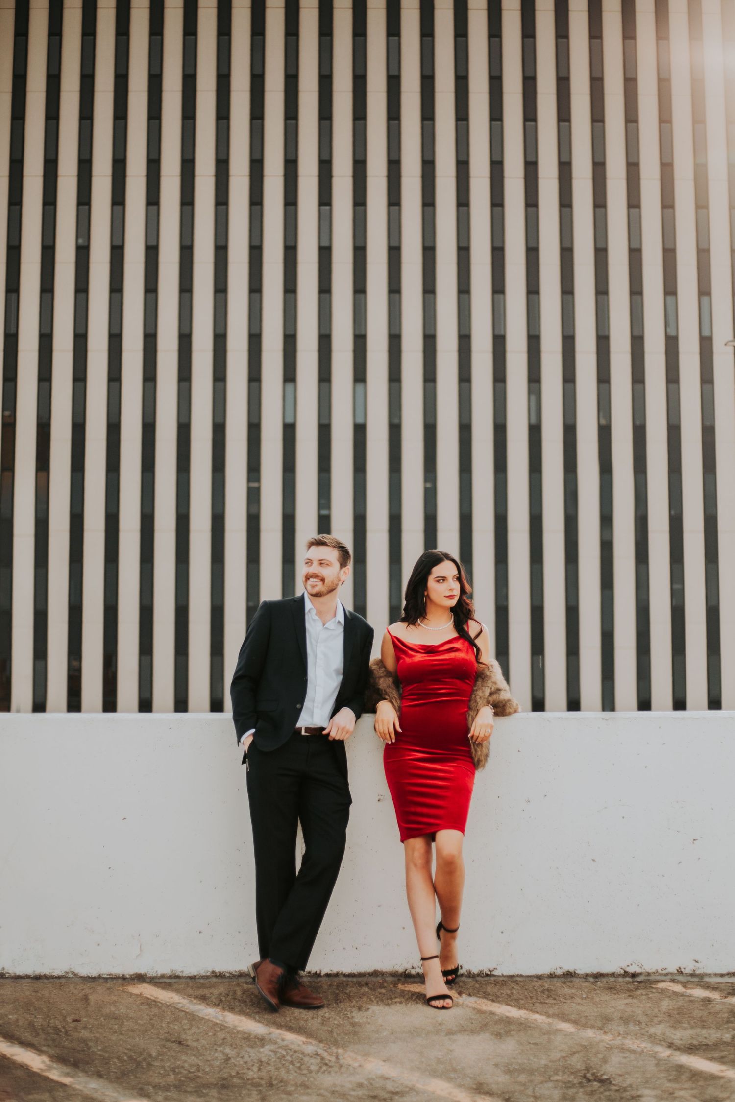 A couple poses against a striped wall, man in black suit, woman in red dress and heels.