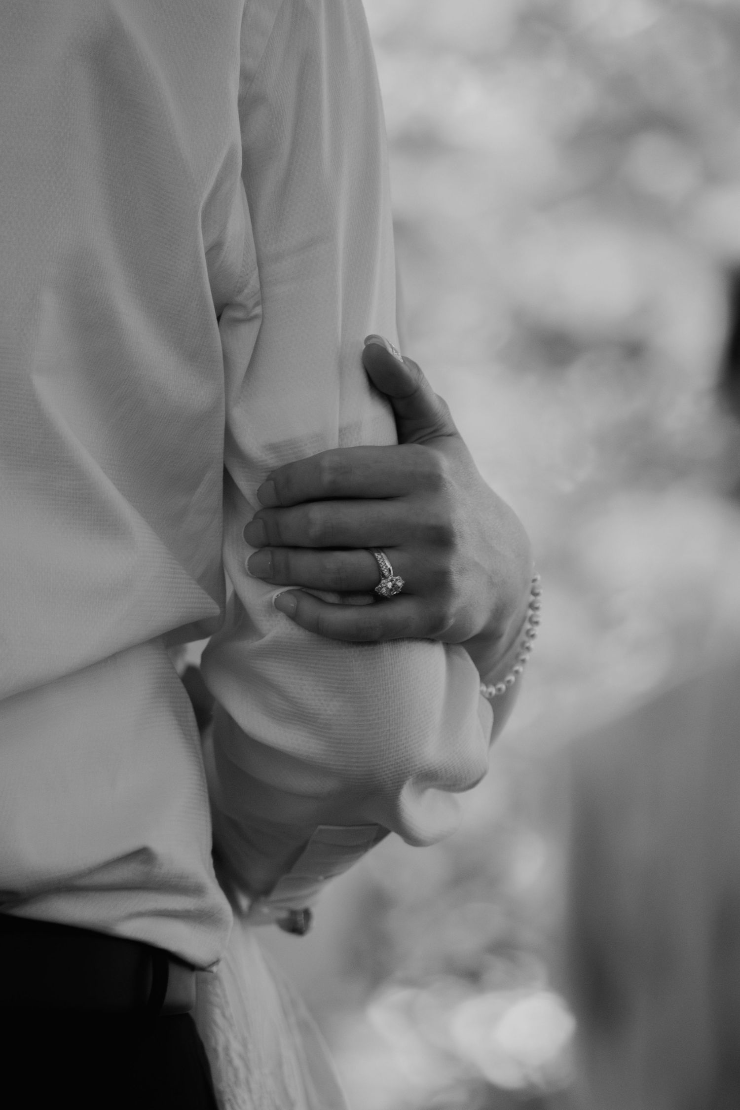 Black and white close-up of bride's hand with diamond engagement ring and pearl bracelet holding groom's arm. Viewpoint at Buckhorn Creek.
