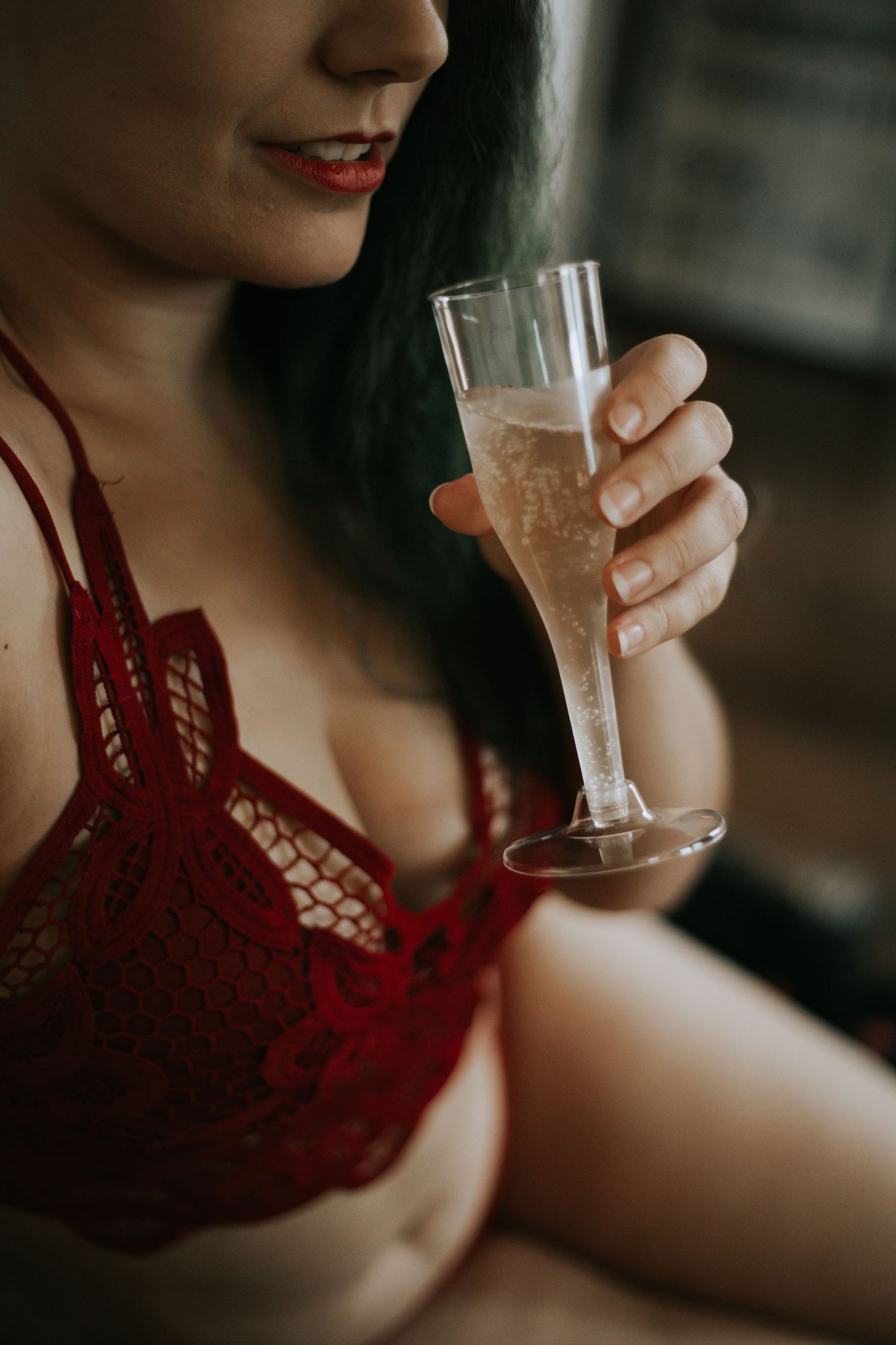 A smiling woman in red lace lingerie holds a champagne flute in a dimly lit intimate setting.