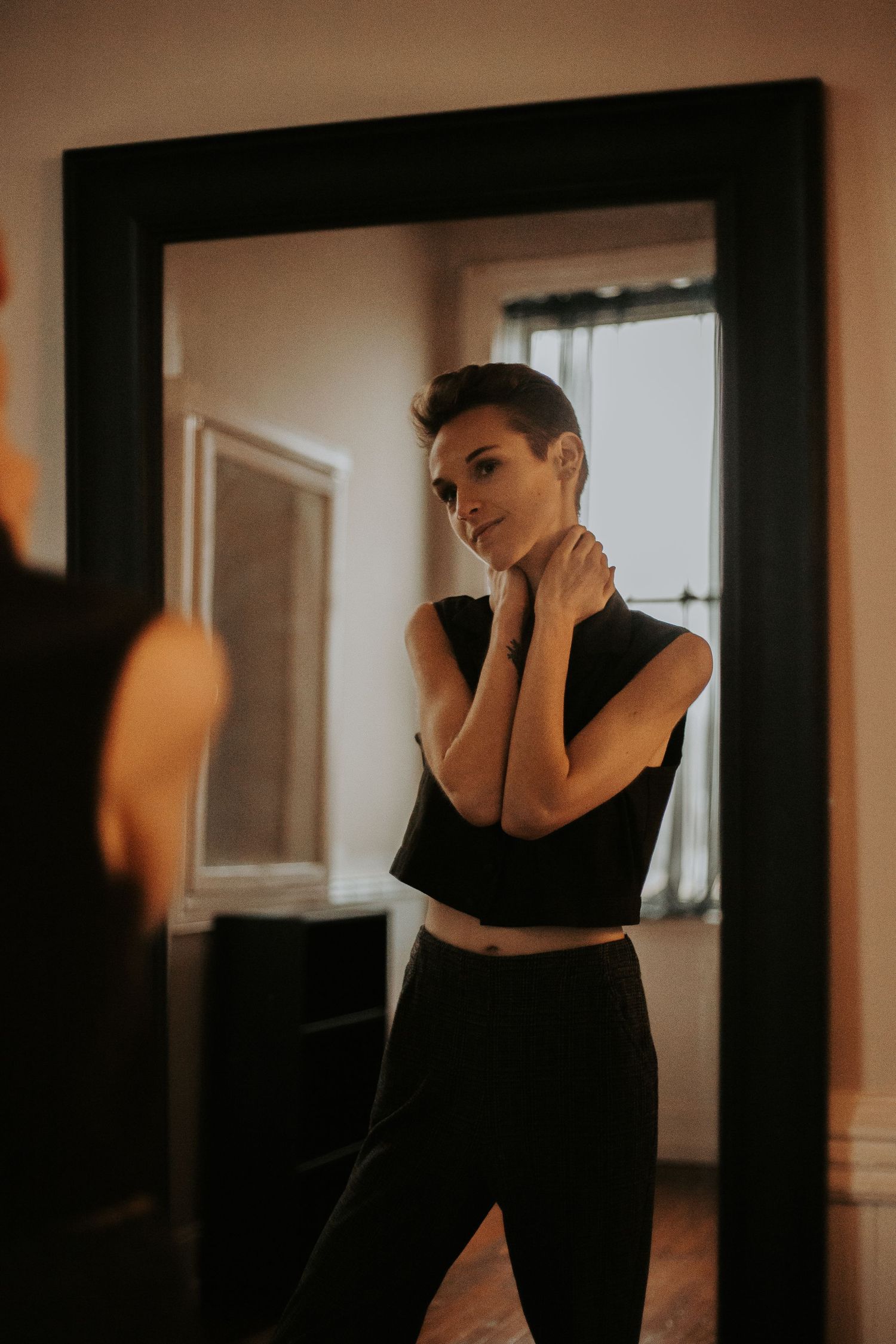 A short-haired woman in a black crop top adjusts her necklace while gazing into a large mirror.. wanderlust boudoir studio honea path sc