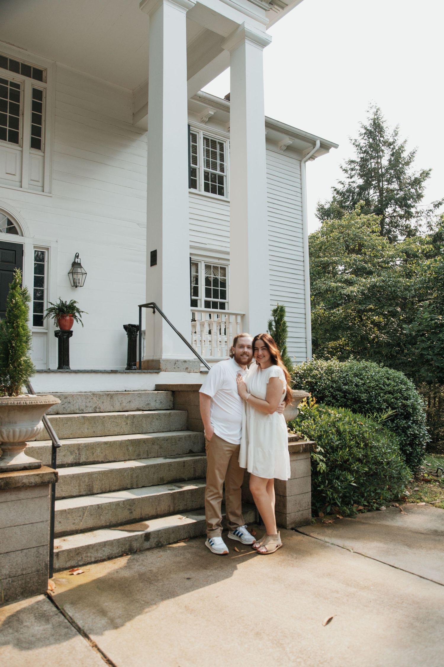 Couple in white outfits posing on steps of elegant white colonial mansion with large pillars and lush greenery.