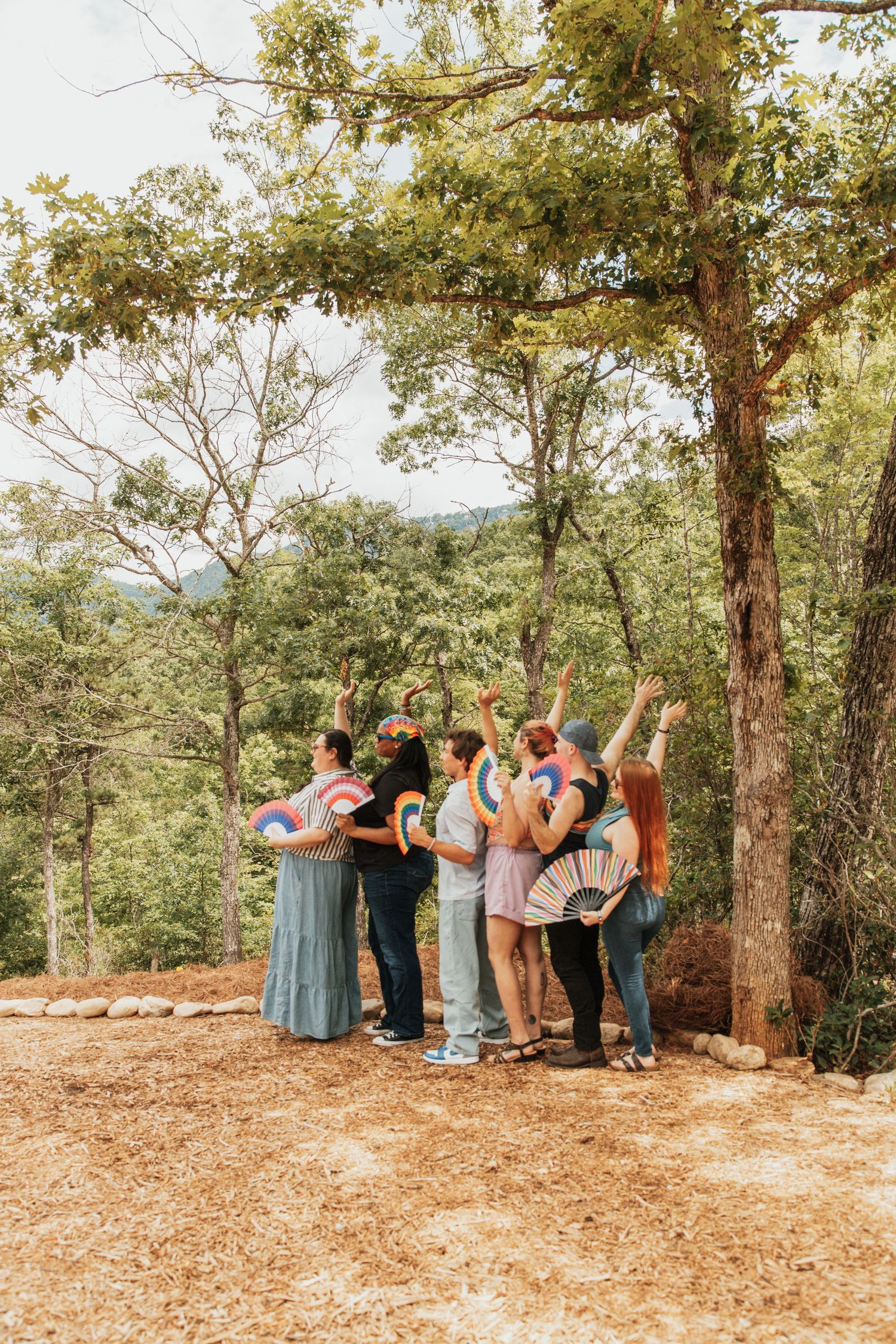 Group of friends holding rainbow fans celebrating outdoors in a lush forest setting on a sunny day.