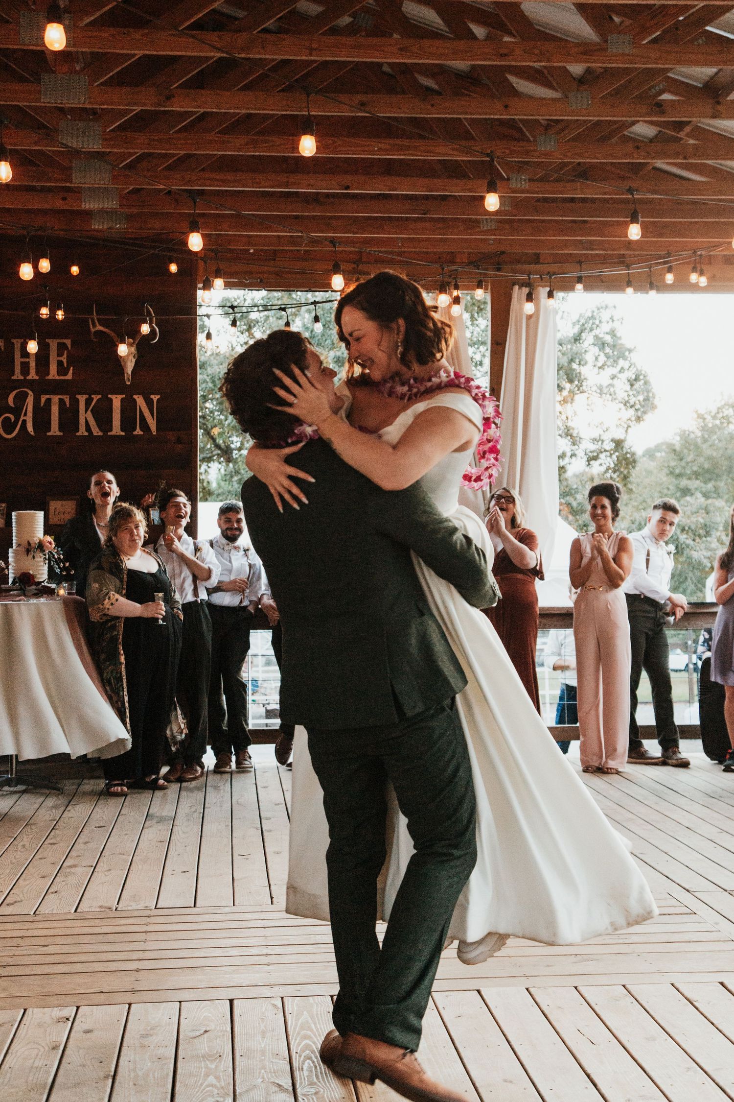 Groom lifts bride during first dance at rustic barn wedding reception with string lights and guests watching.