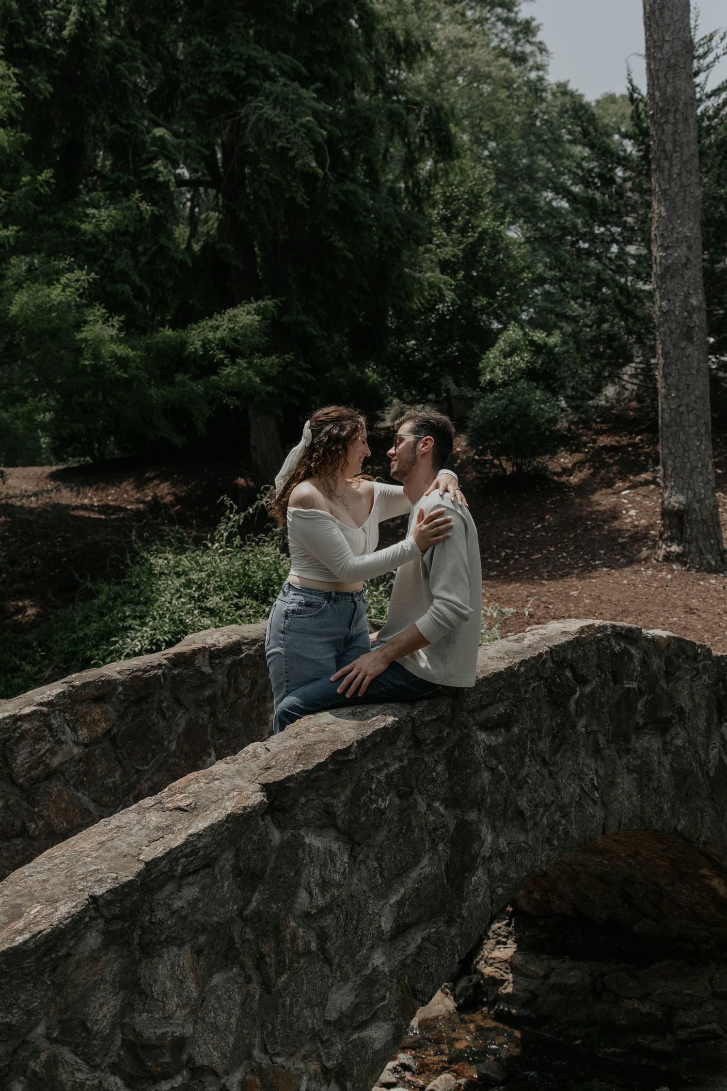 Couple sitting on a stone bridge in a lush forest, sharing a romantic moment while facing each other.