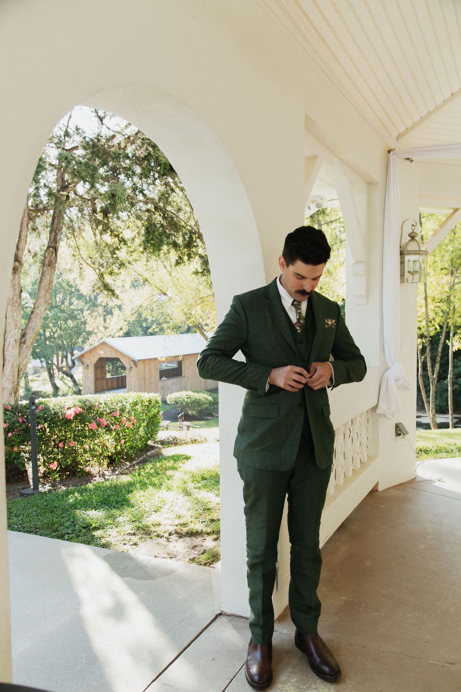 Groom in forest green three-piece suit buttoning jacket on sunlit white porch with garden backdrop. venue at ballenger bridge landrum sc