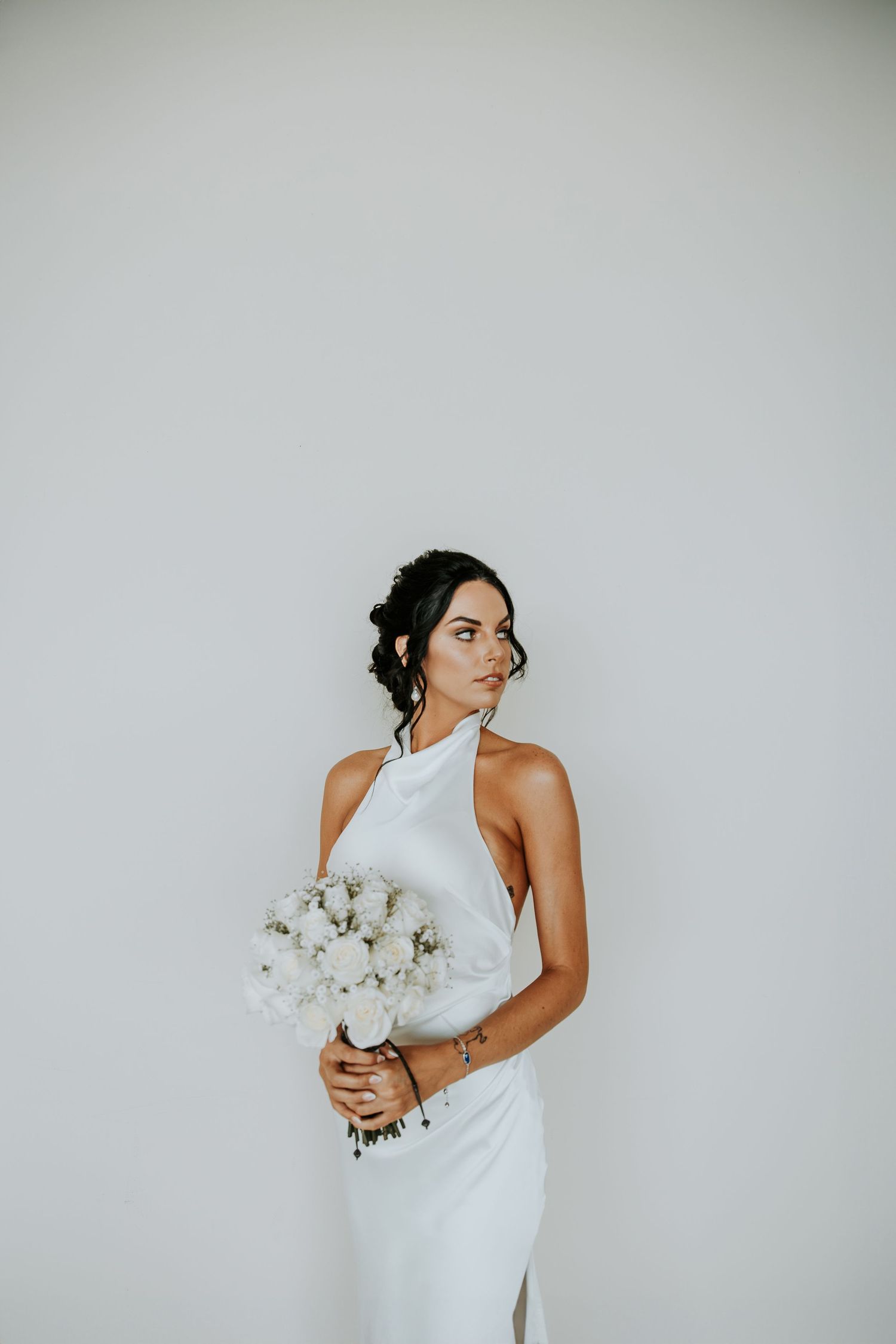Elegant bride in white halter gown holds white floral bouquet against white wall. - riviera maya resort (cancun, Mexico)