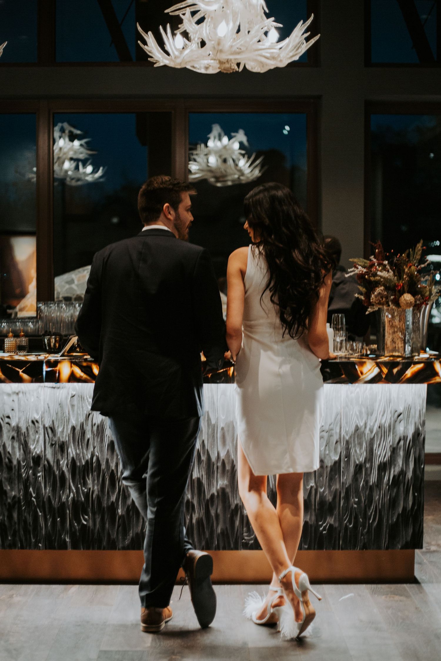 Couple in formal attire standing at an elegant bar with a white chandelier and decorative backdrop at a nighttime event.