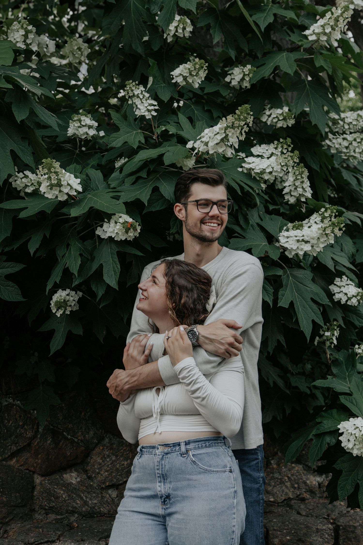 Happy couple embracing in front of lush green bushes with white flowers, smiling outdoors in casual attire.