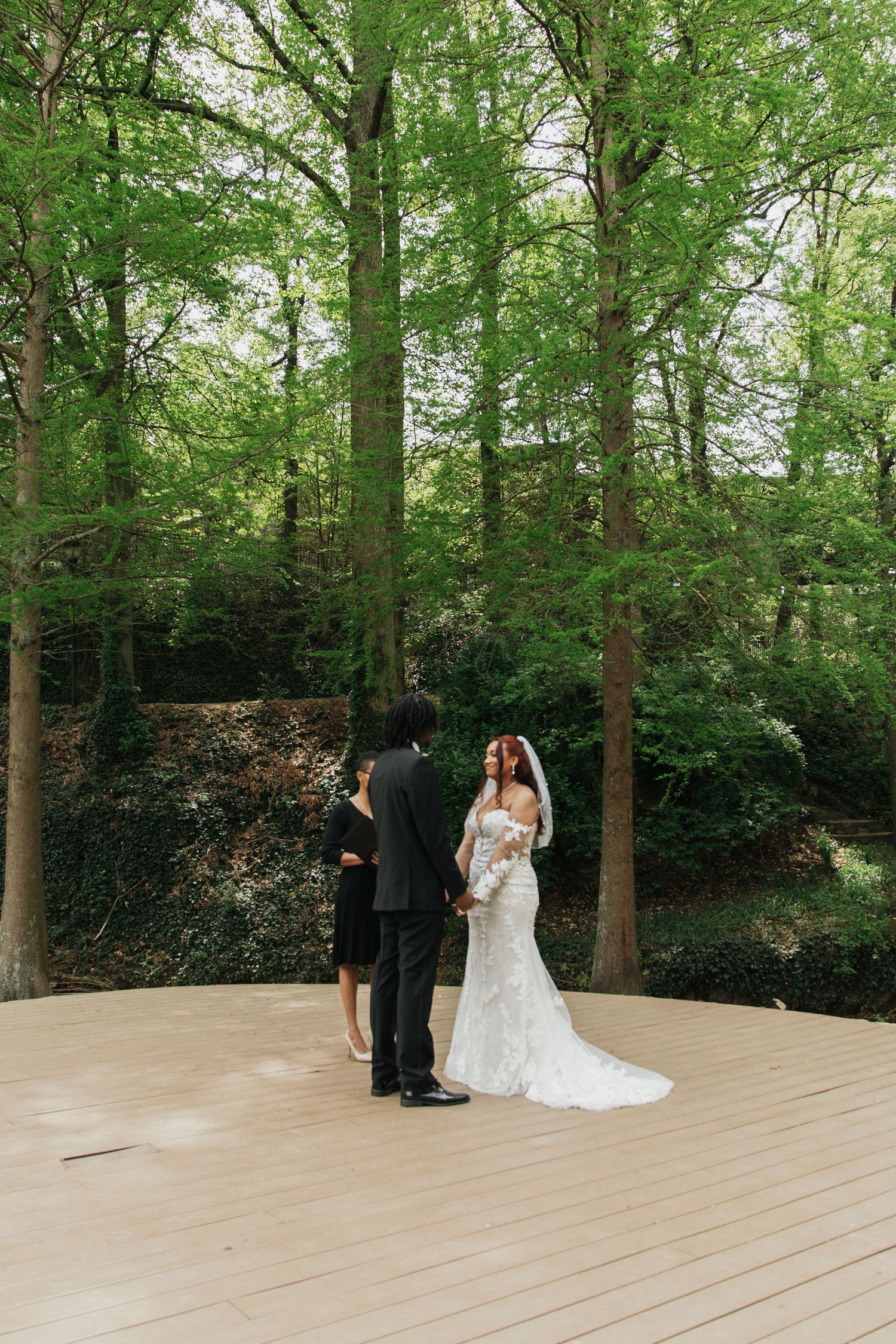 Bride in lace gown stands on wooden deck platform surrounded by tall green trees.  falls park greenville sc