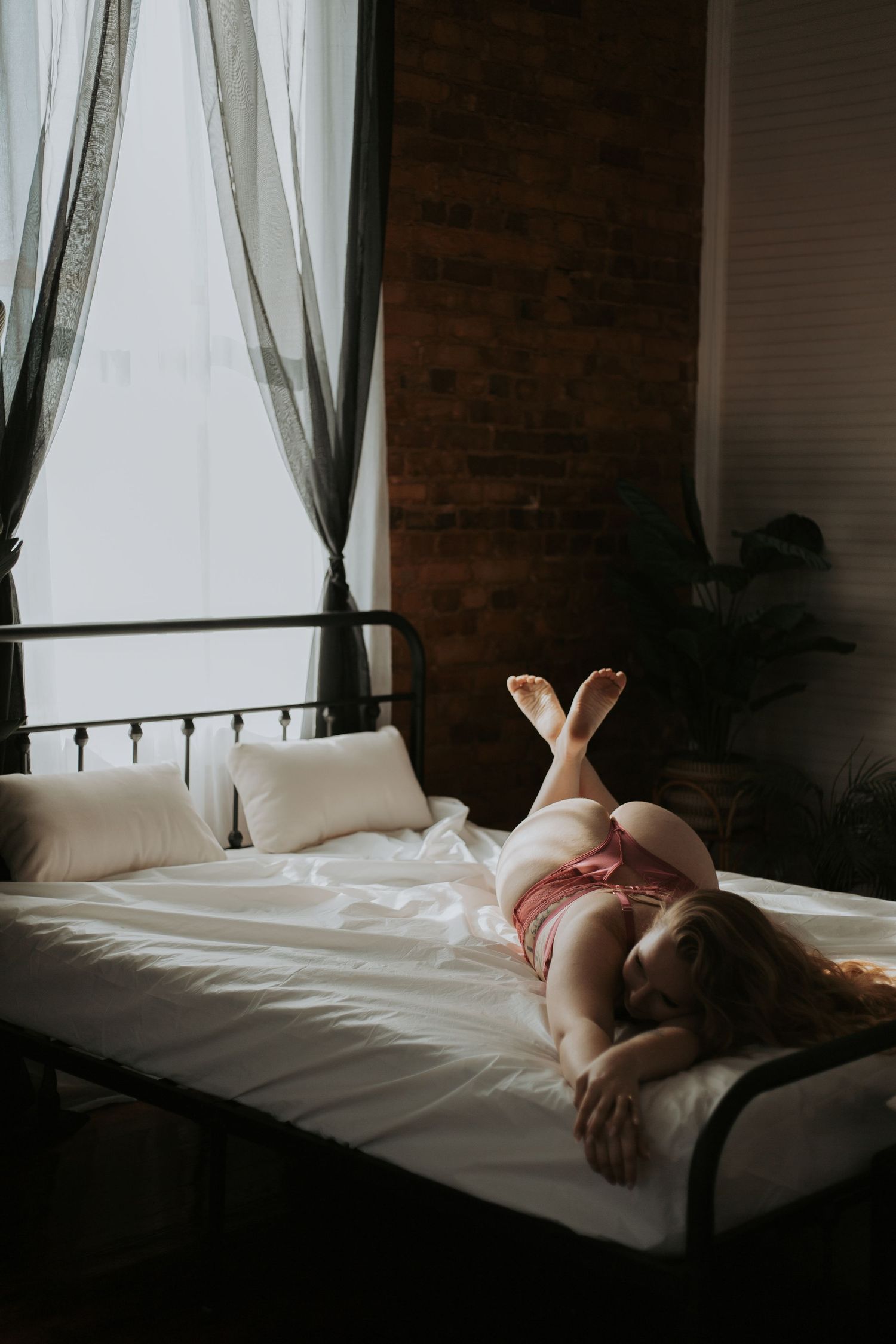 Woman in pink lingerie lying on a white bed near a large window in a brick loft.. wanderlust boudoir studio honea path sc
