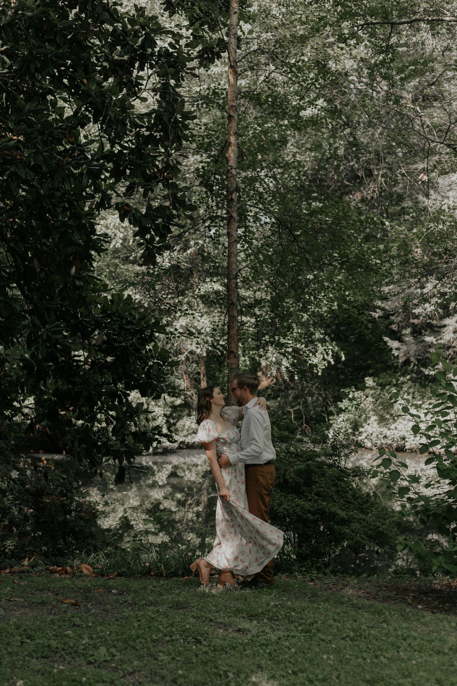 Couple dancing romantically under lush green trees in a garden, woman wearing a flowing white lace dress.