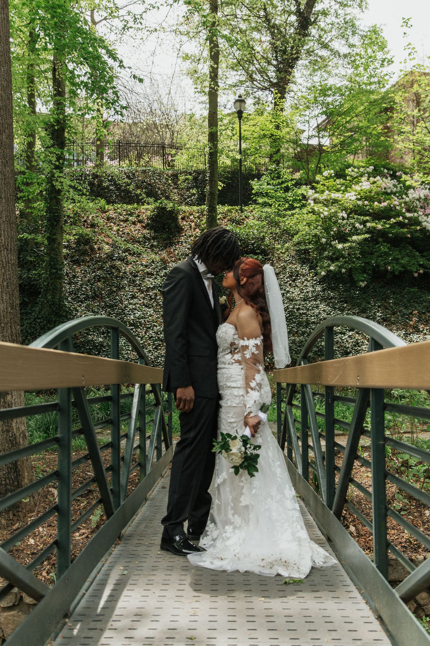 Bride and groom share a kiss on a garden bridge, she in a floral gown with veil, he in a black suit.