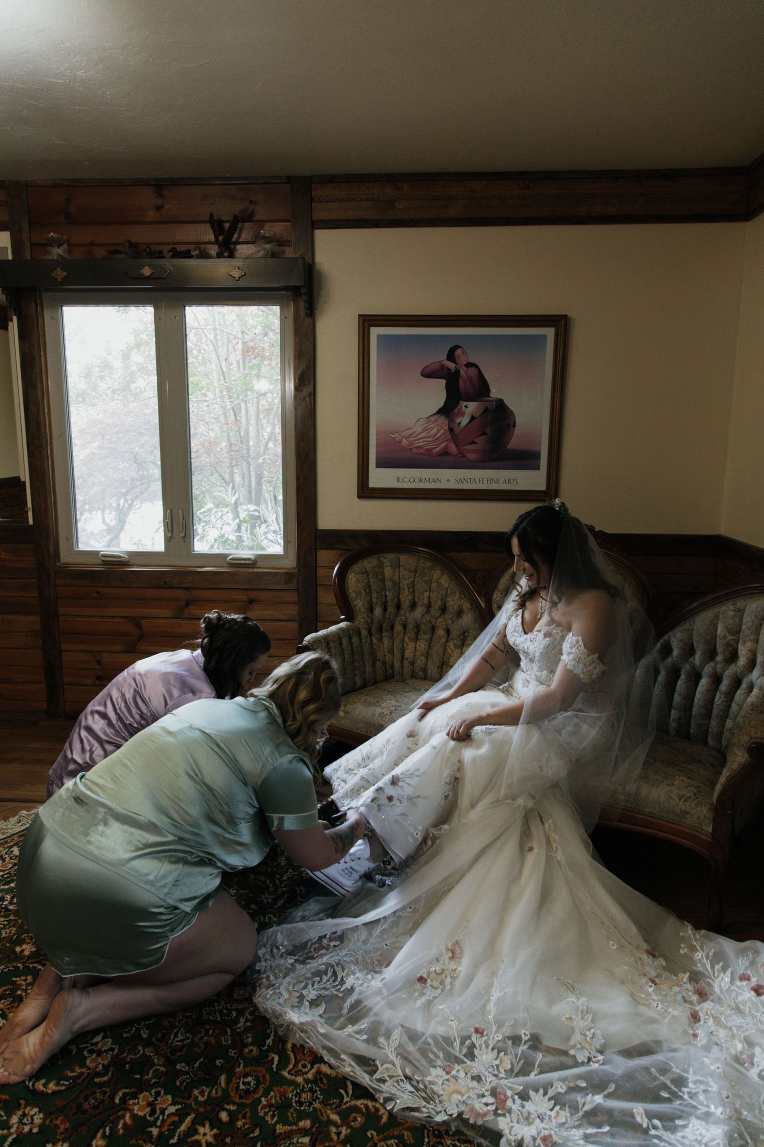 Bride in lace wedding gown being helped by bridesmaid in rustic wood-paneled room before wedding ceremony.
