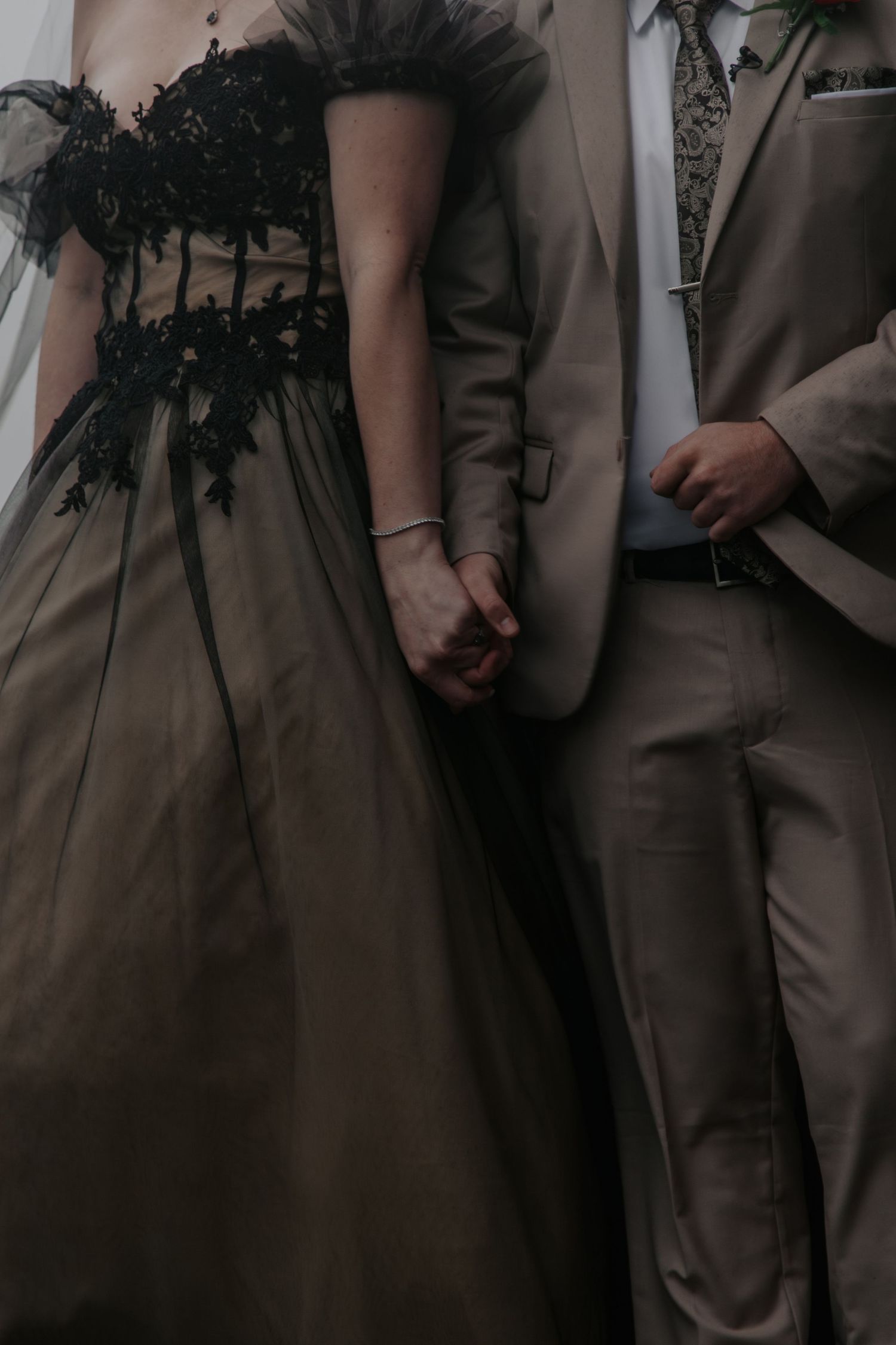 Close-up of bride and groom holding hands, bride in black lace corset olive tulle gown, groom in tan suit.
