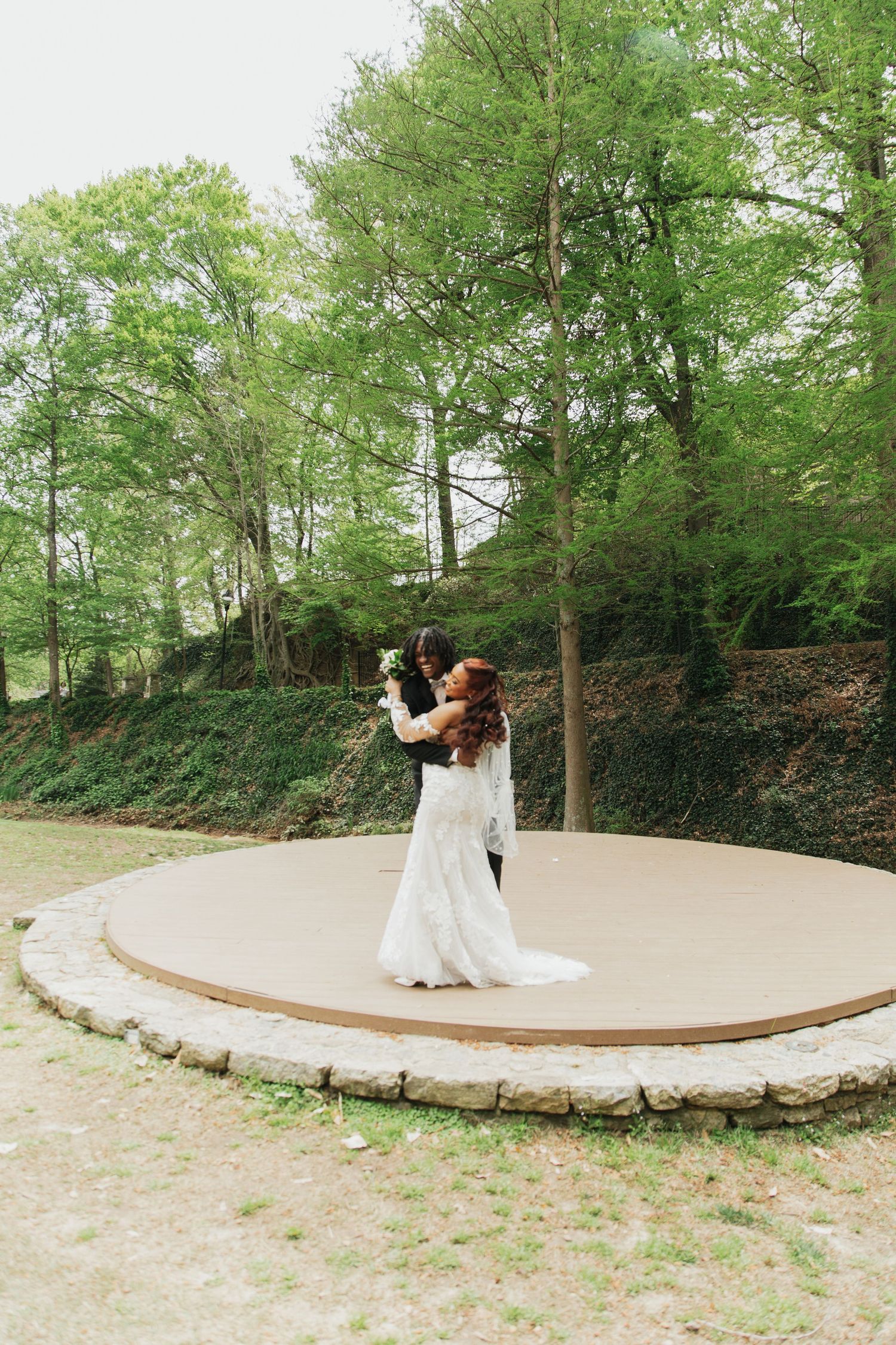 Bride and groom embrace on circular wooden dance platform surrounded by lush green trees.  falls park greenville sc